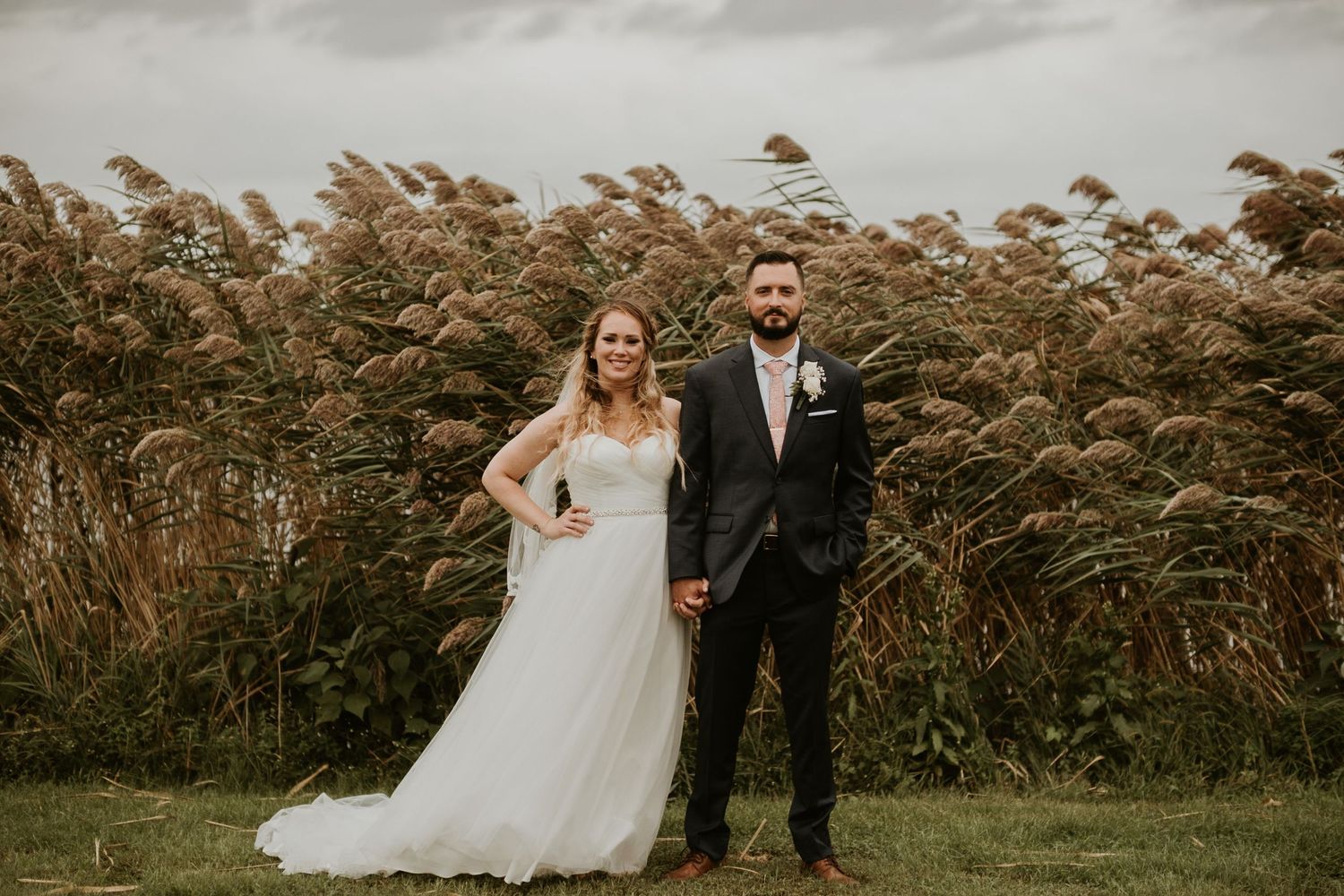 Bride and groom holding hands in front of tall golden reeds on a cloudy day at an outdoor wedding venue.
