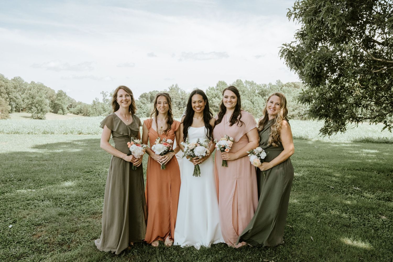 A group in formal attire poses outdoors in a garden setting with coordinated earth-toned dresses and floral bouquets.