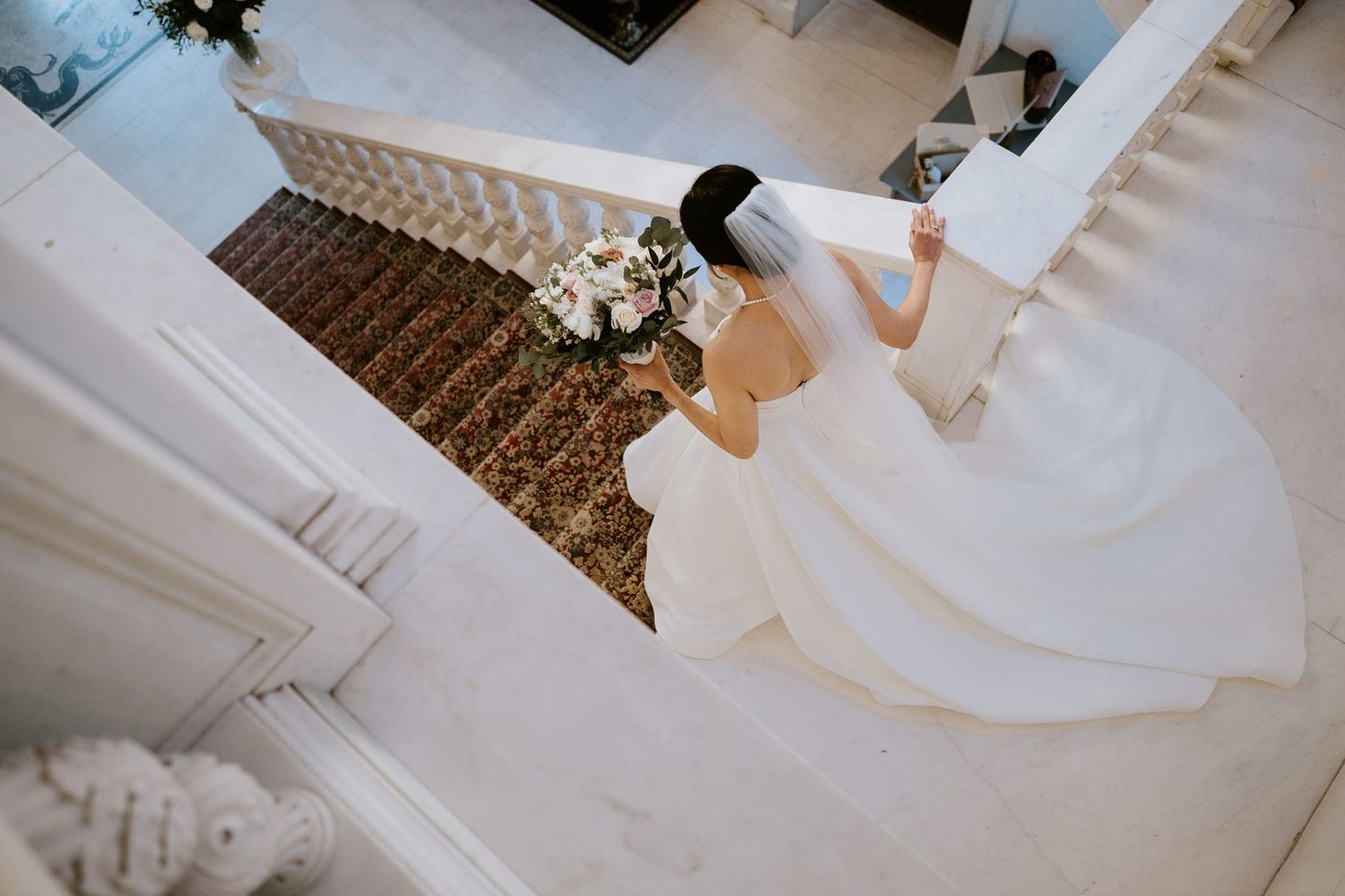 Bride in elegant white gown descending grand marble staircase holding bouquet, aerial view wedding photography.