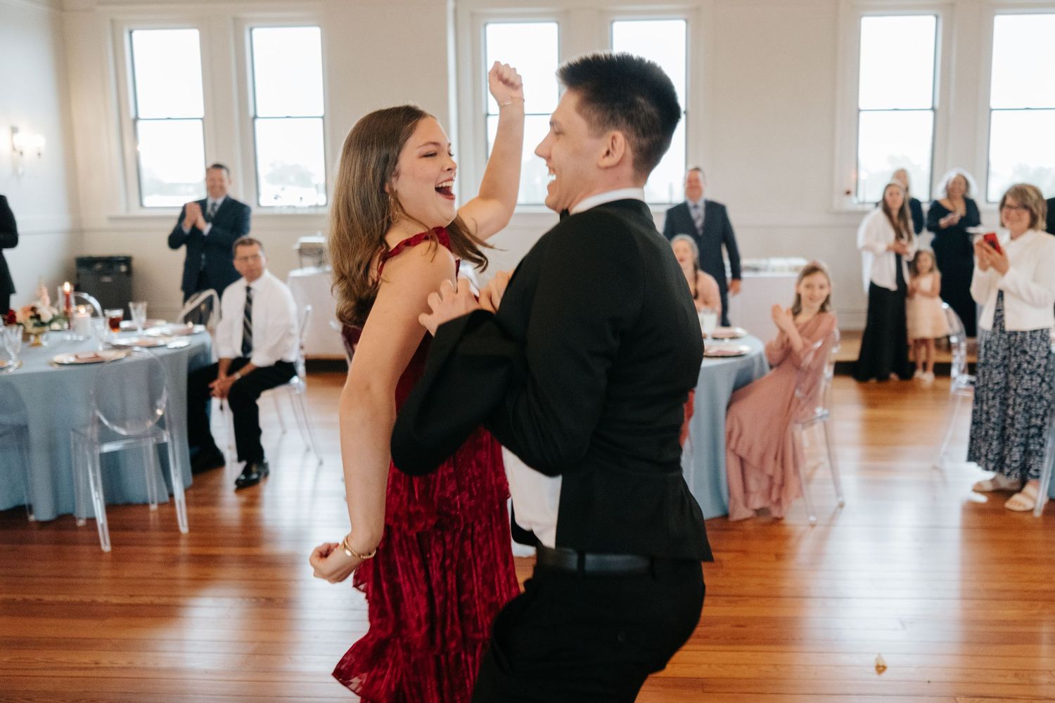 Wedding reception dance floor scene showing couples dancing in an elegant white-walled venue with windows.