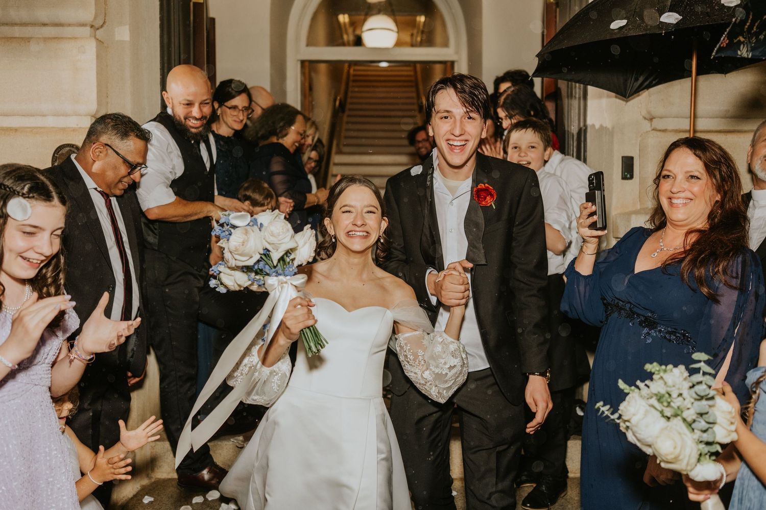 A joyous wedding couple exits a building surrounded by celebrating guests throwing confetti and holding sparklers.