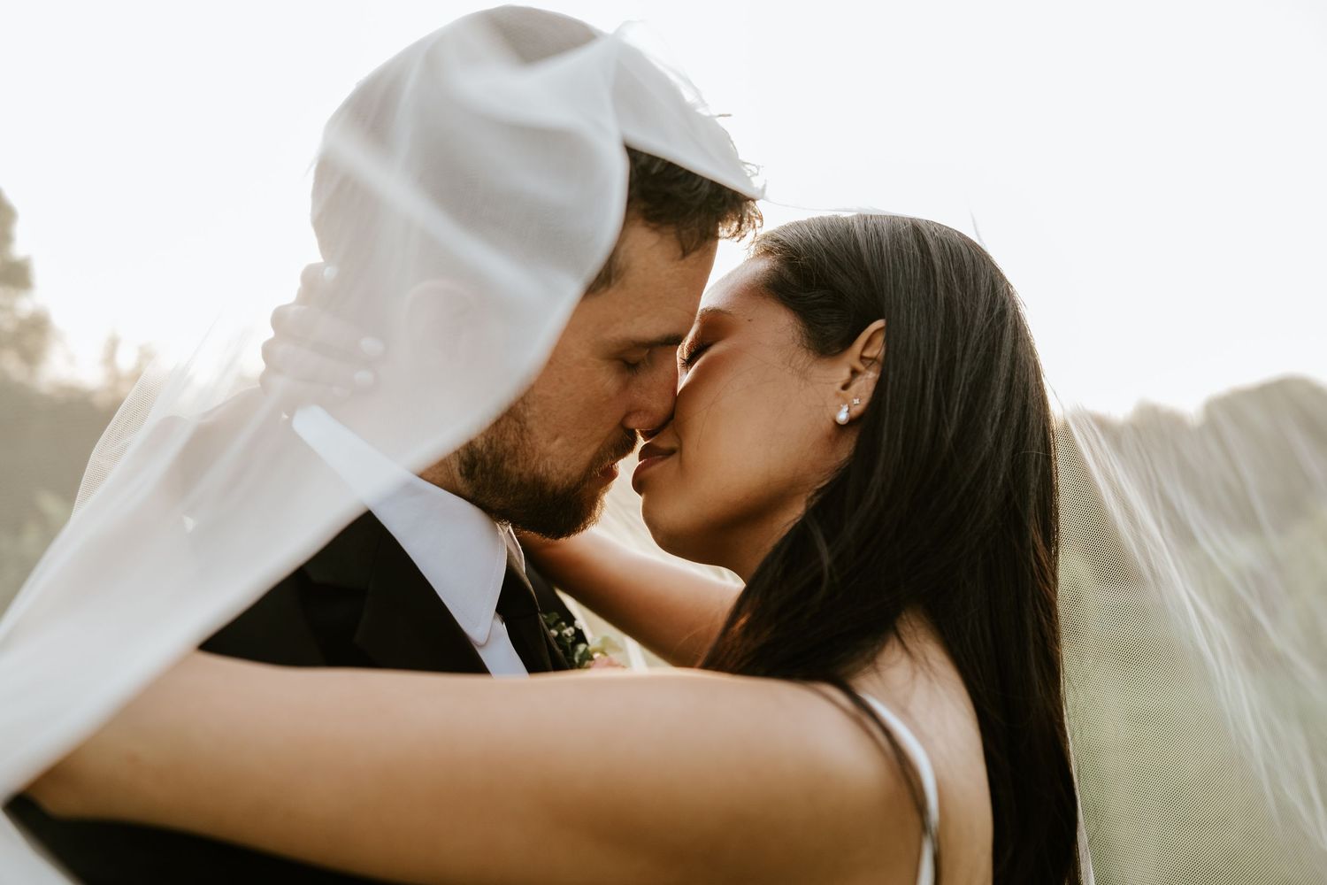 A romantic couple shares a tender kiss while white fabric billows around them in soft natural light.
