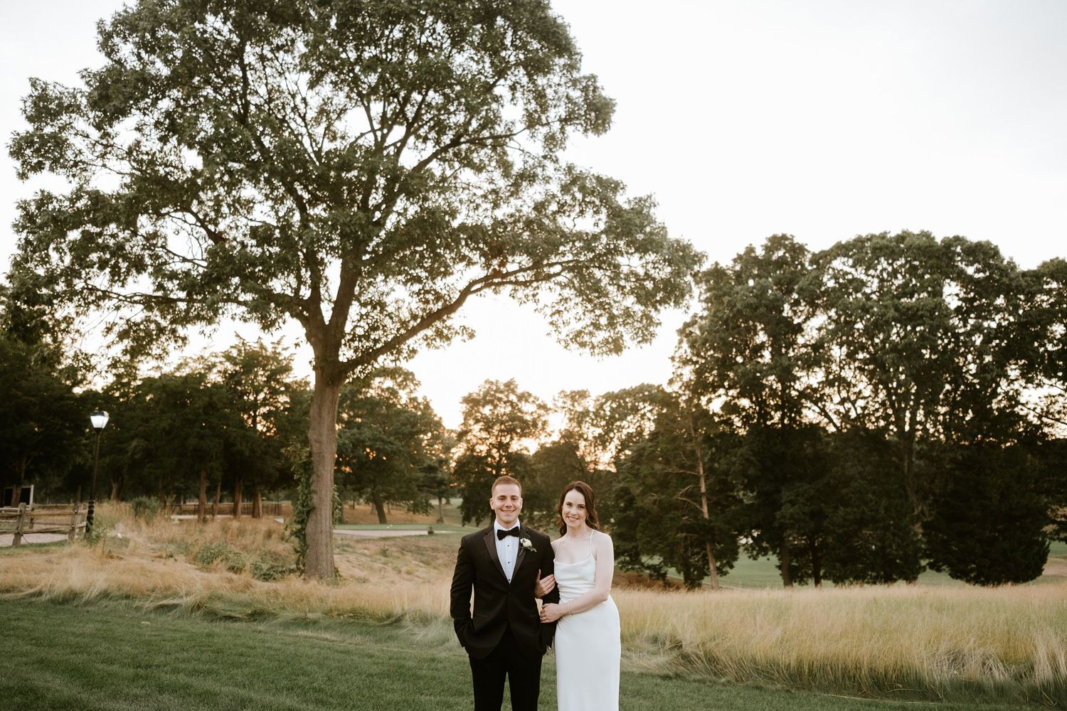 Bride and groom standing together on lush green lawn at sunset with tall oak trees in background at outdoor wedding.