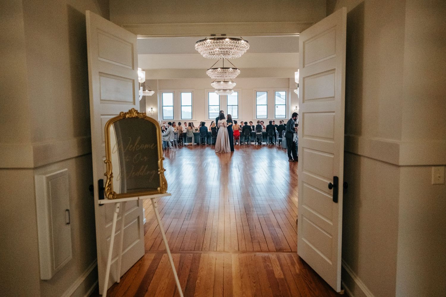 Long hallway with white wainscoting and hardwood floors leads to bright windows in an elegant venue.