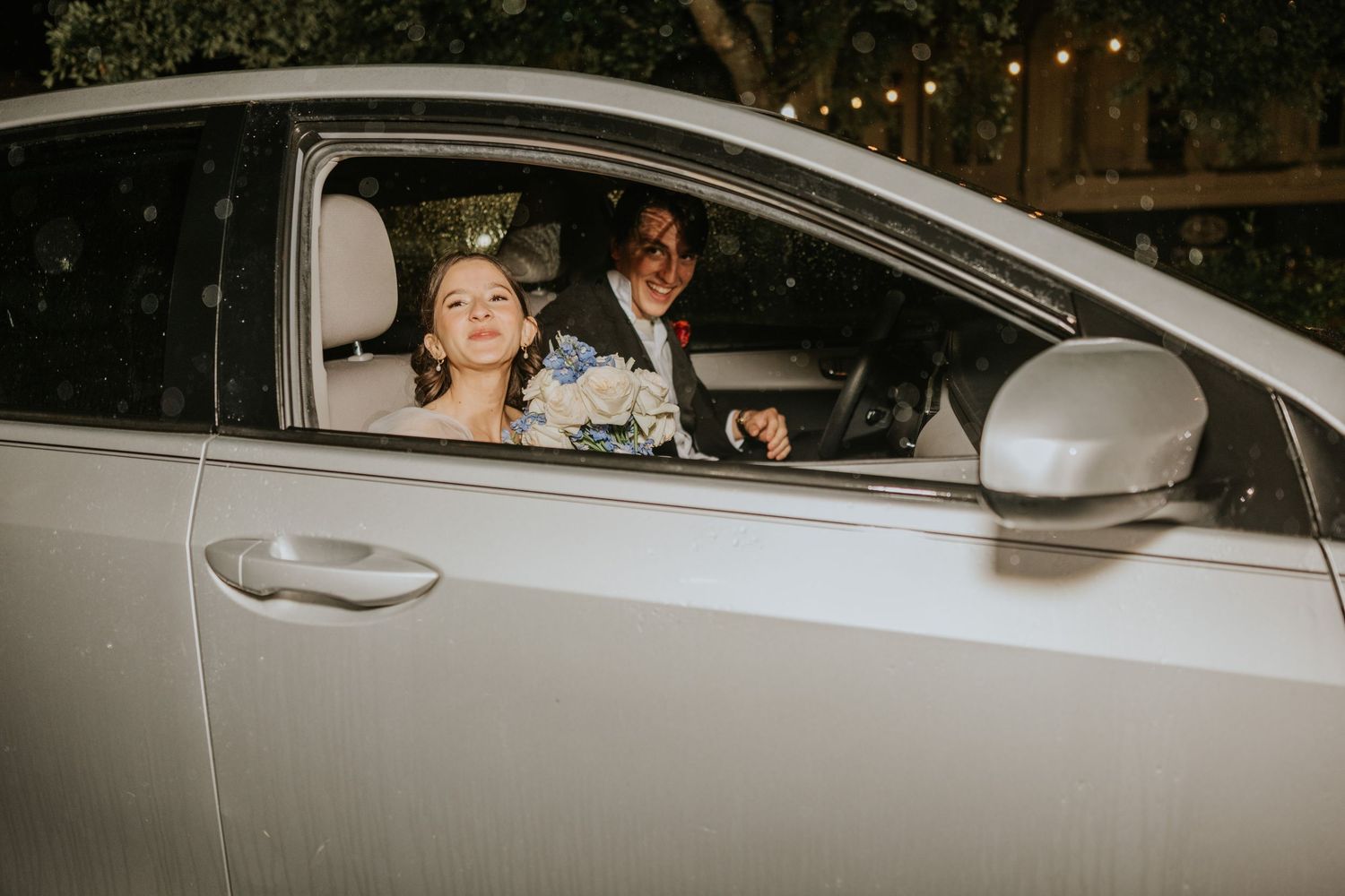 A couple in wedding attire sits in a white car at night, with string lights visible in the background.