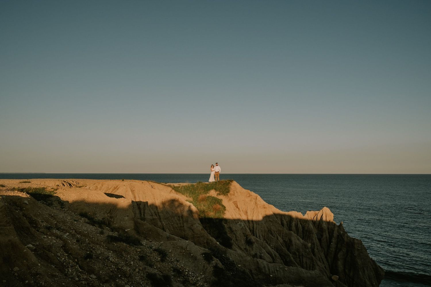 Silhouettes of two people stand on rocky cliffs against a serene ocean backdrop at sunset.
