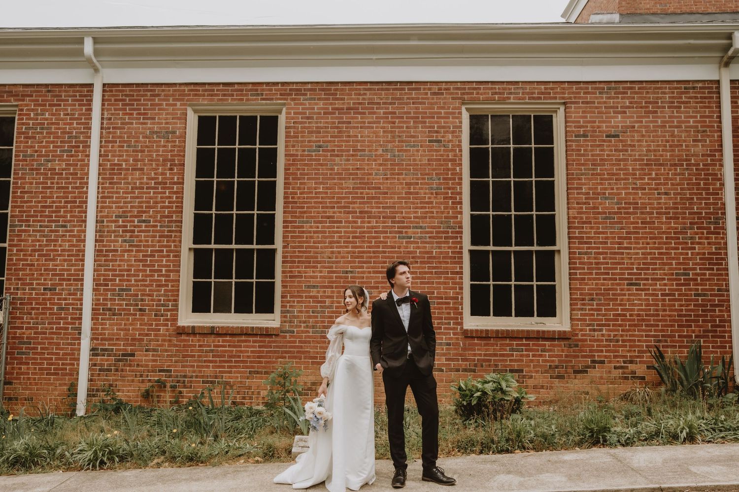 A couple poses together against a red brick building exterior with white-trimmed windows.
