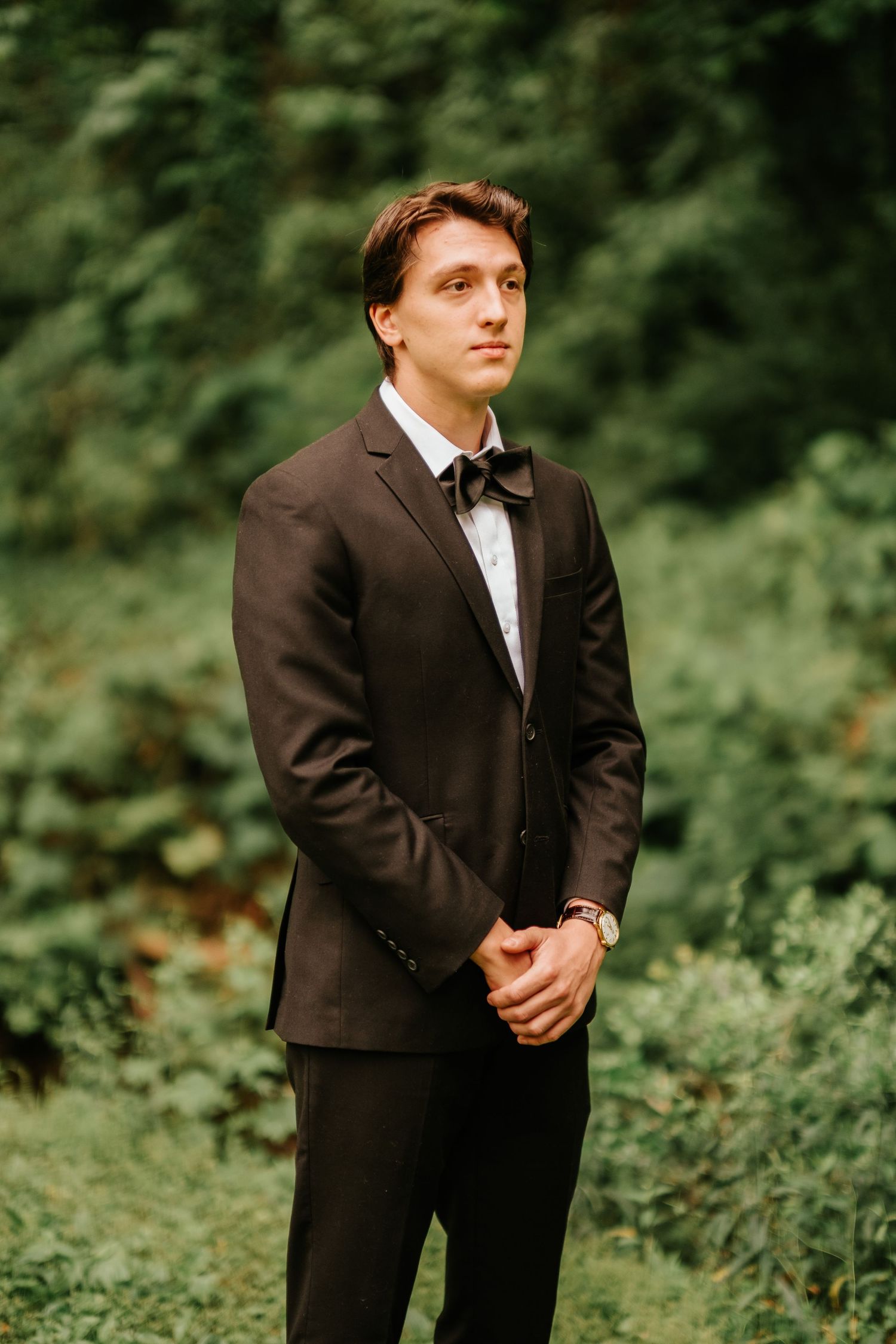 Groom in a black tuxedo poses against a green natural backdrop in an outdoor setting.