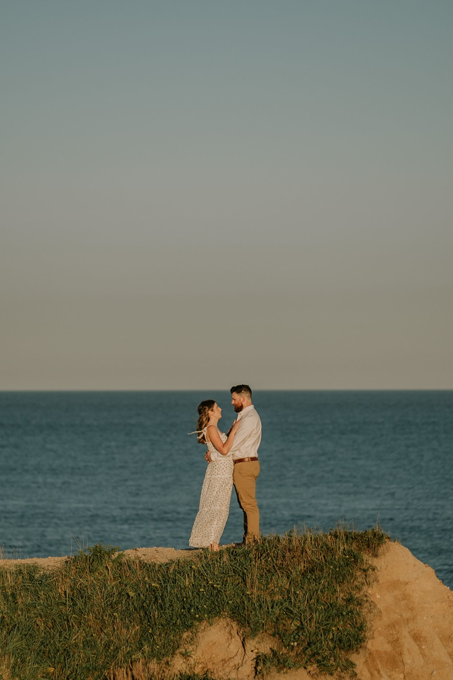 A couple stands together on a coastal cliff overlooking the ocean during golden hour.