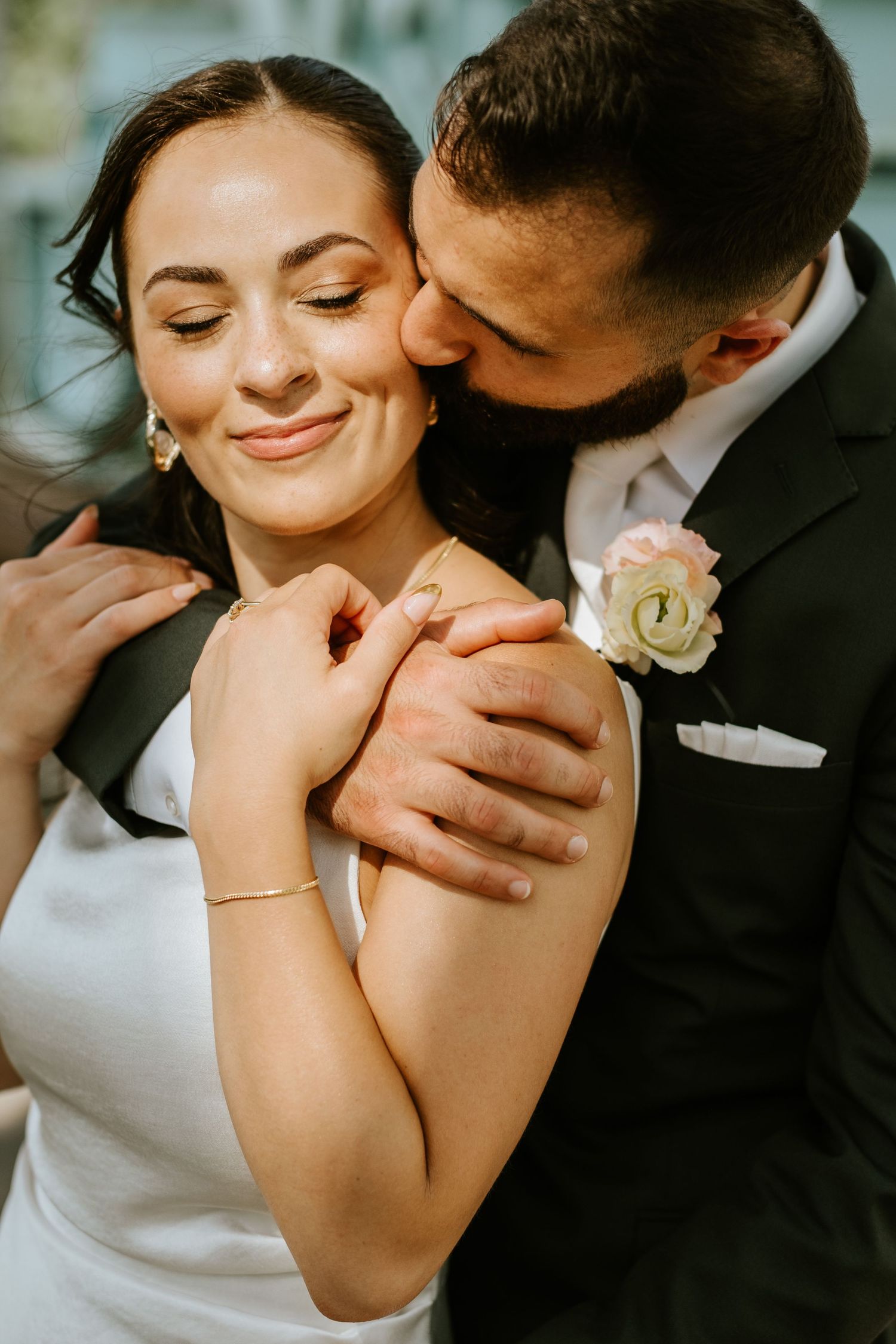 Groom in black suit kissing bride on cheek, bride smiling with eyes closed, wearing white dress and gold bracelet.