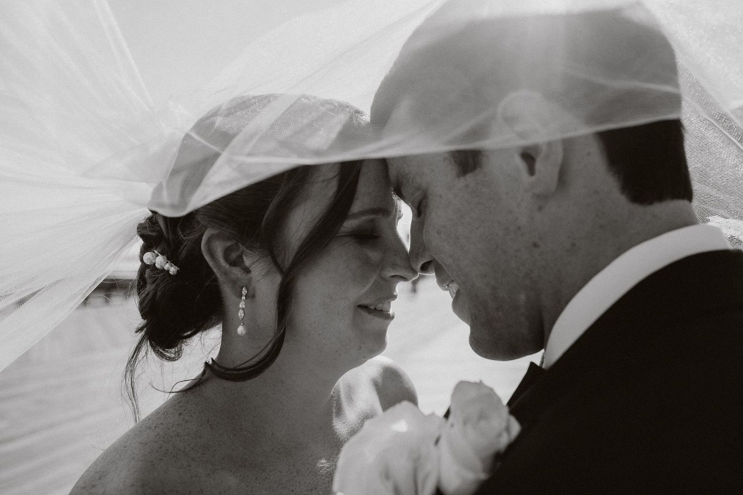 Black and white wedding photo of bride and groom touching foreheads under a flowing veil.
