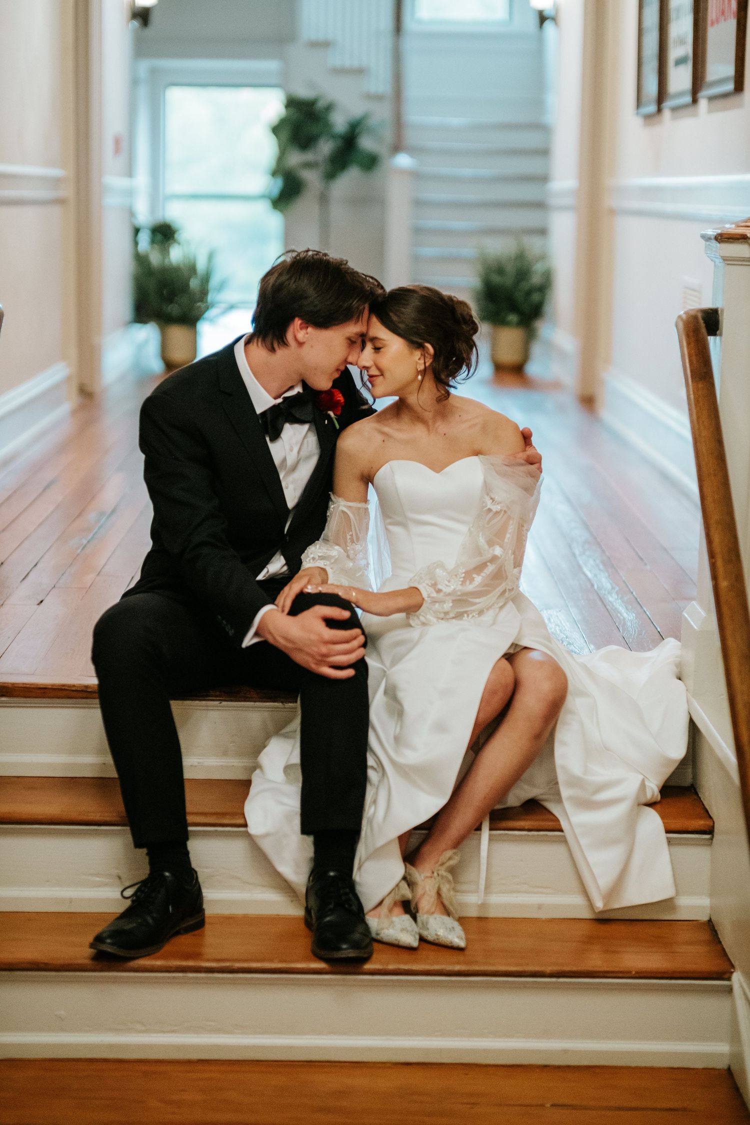 A couple in wedding attire sits together on wooden floor boards in an elegant white interior space.