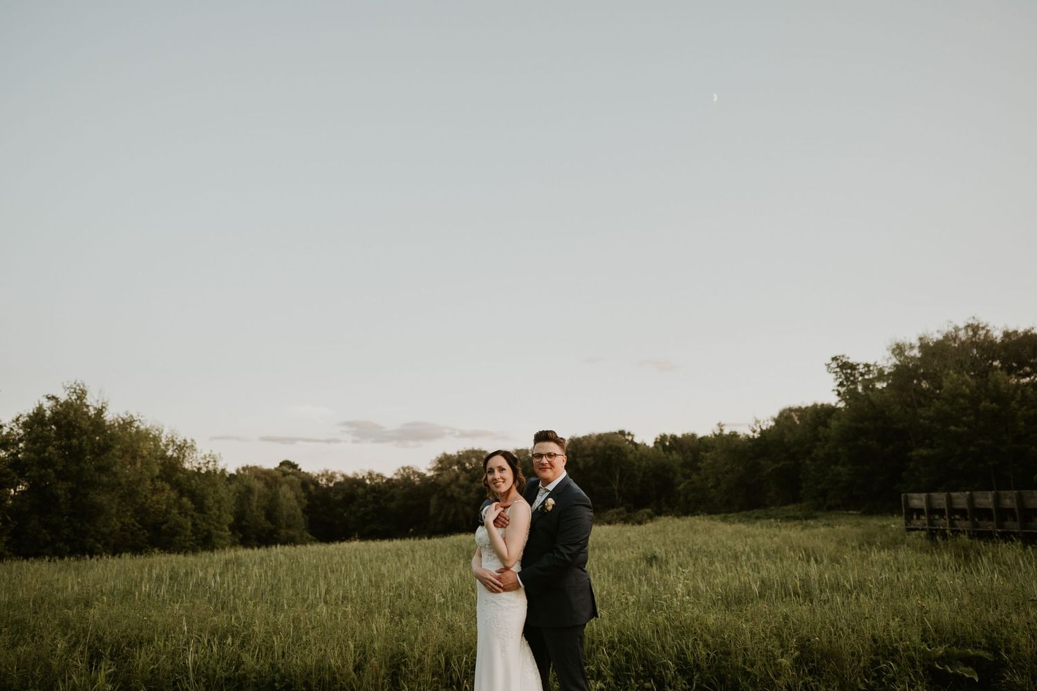 A bride and groom pose together in a lush green field at dusk, surrounded by trees in a romantic outdoor wedding portrait.