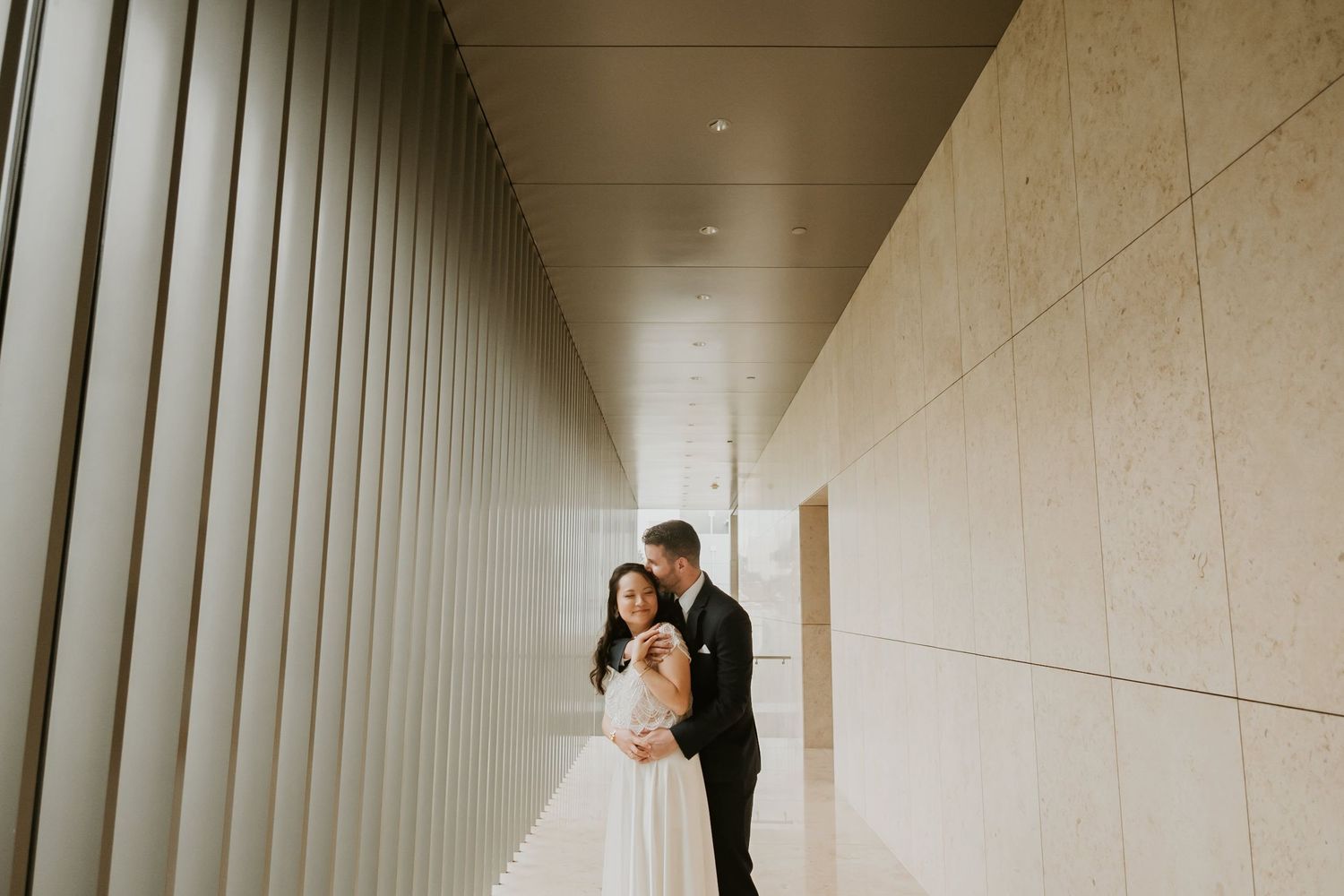 Bride and groom embracing in a modern architectural hallway with warm stone walls on their wedding day.