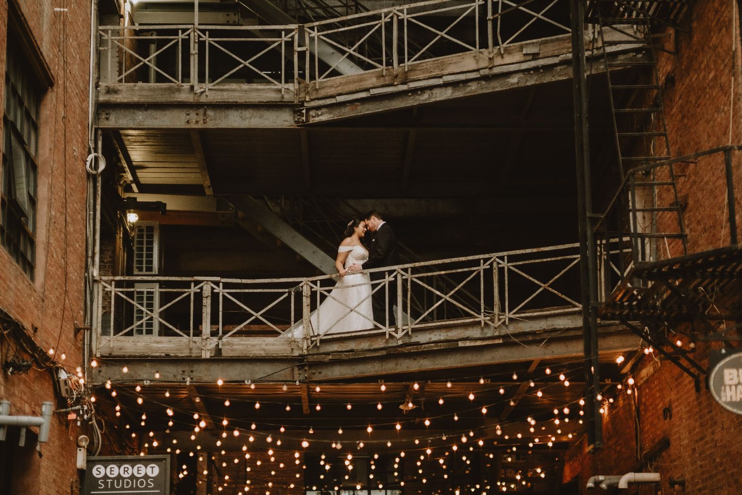 Bride and groom share a romantic kiss on a metal balcony adorned with warm string lights at an industrial wedding venue.