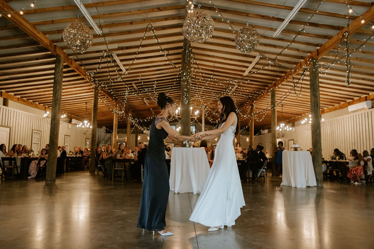 A couple shares their first dance in a rustic barn venue decorated with string lights and surrounded by wedding guests.