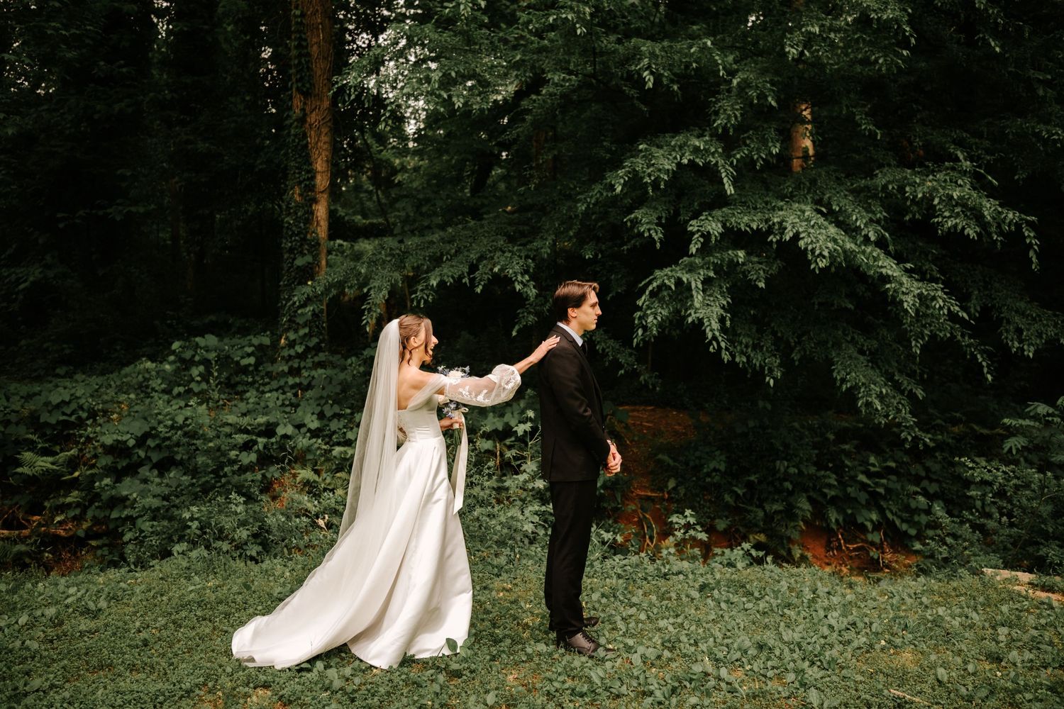 Bride walks to his soon-to-be groom for their first look through a lush forest garden filled with tall evergreen trees.