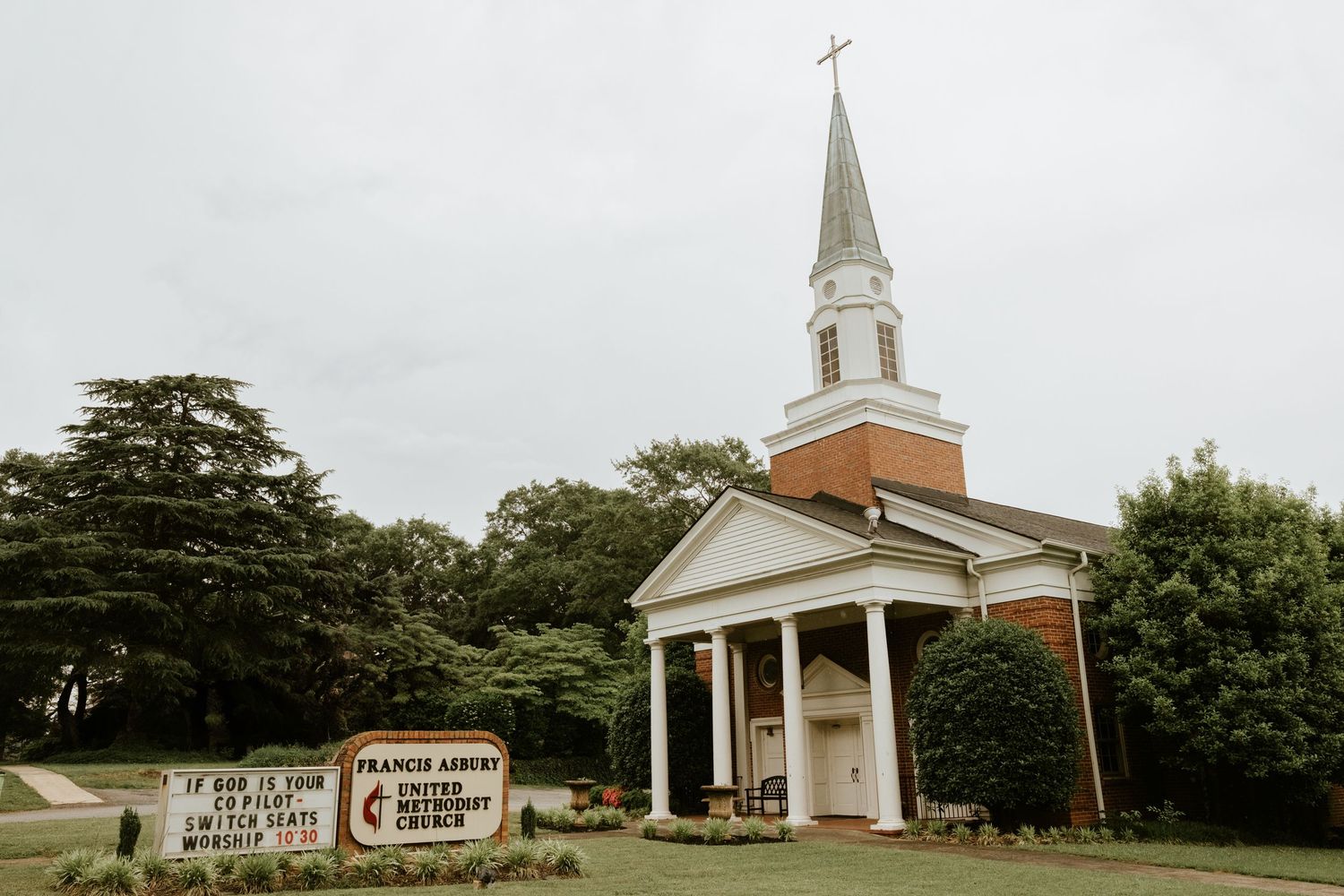 A panoramic view of a traditional white church building with steeple set against a cloudy sky and surrounded by trees.