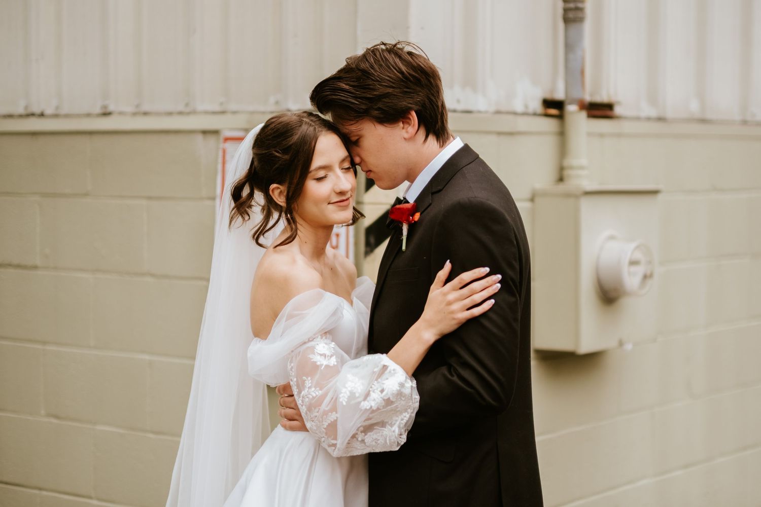 Newlyweds share an intimate moment against a light colored wall in soft natural lighting.