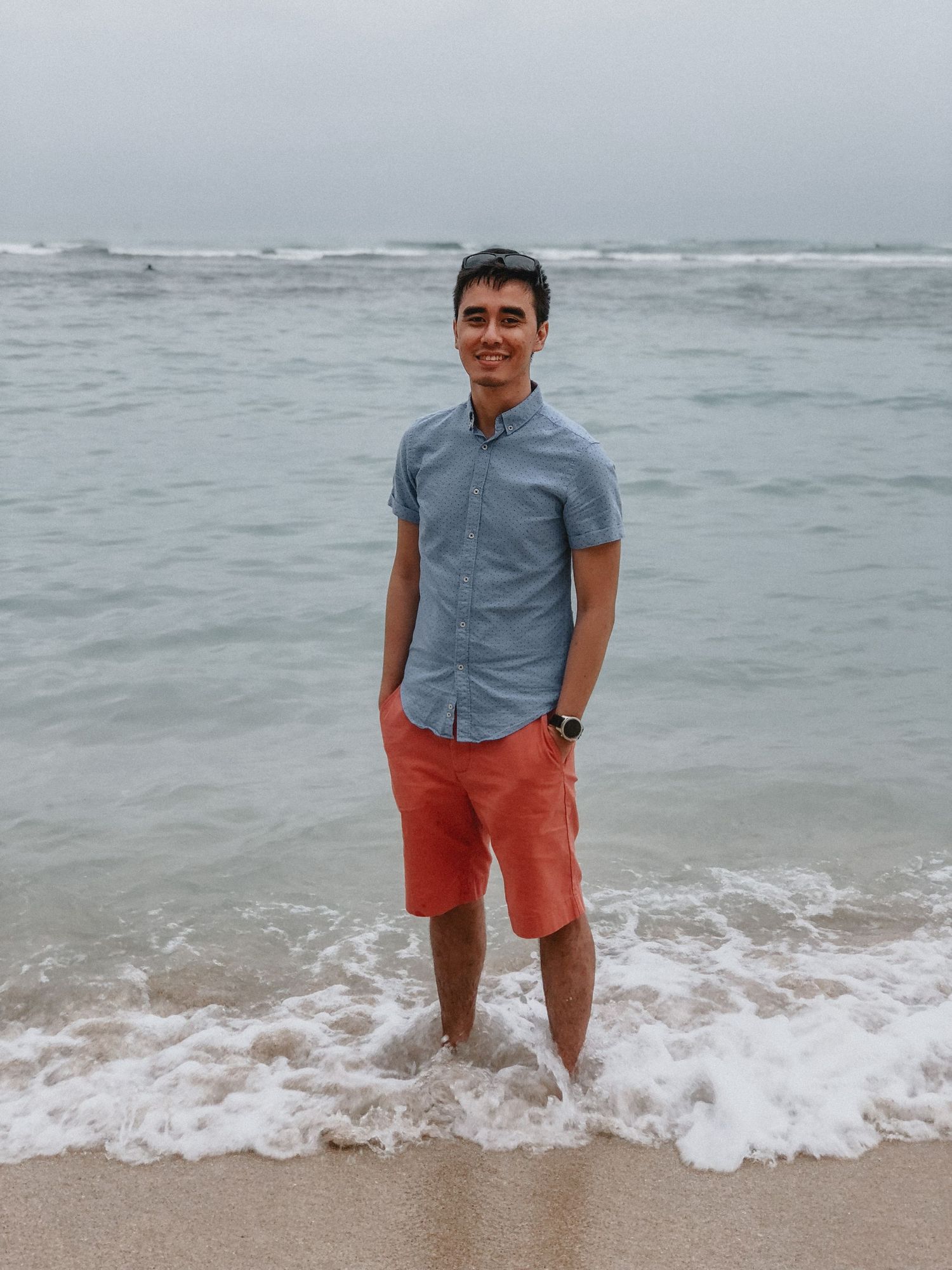 Young man smiling on beach, wearing blue shirt and coral shorts, standing in ocean waves on overcast day.