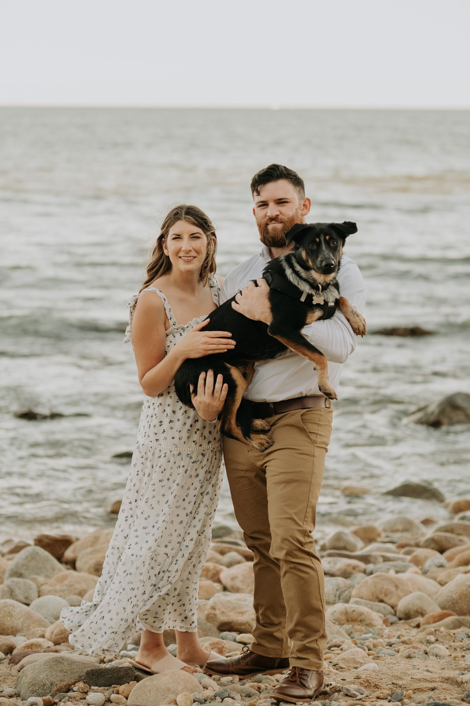 Couple with their black dog poses together on a rocky beach shoreline during an overcast day.