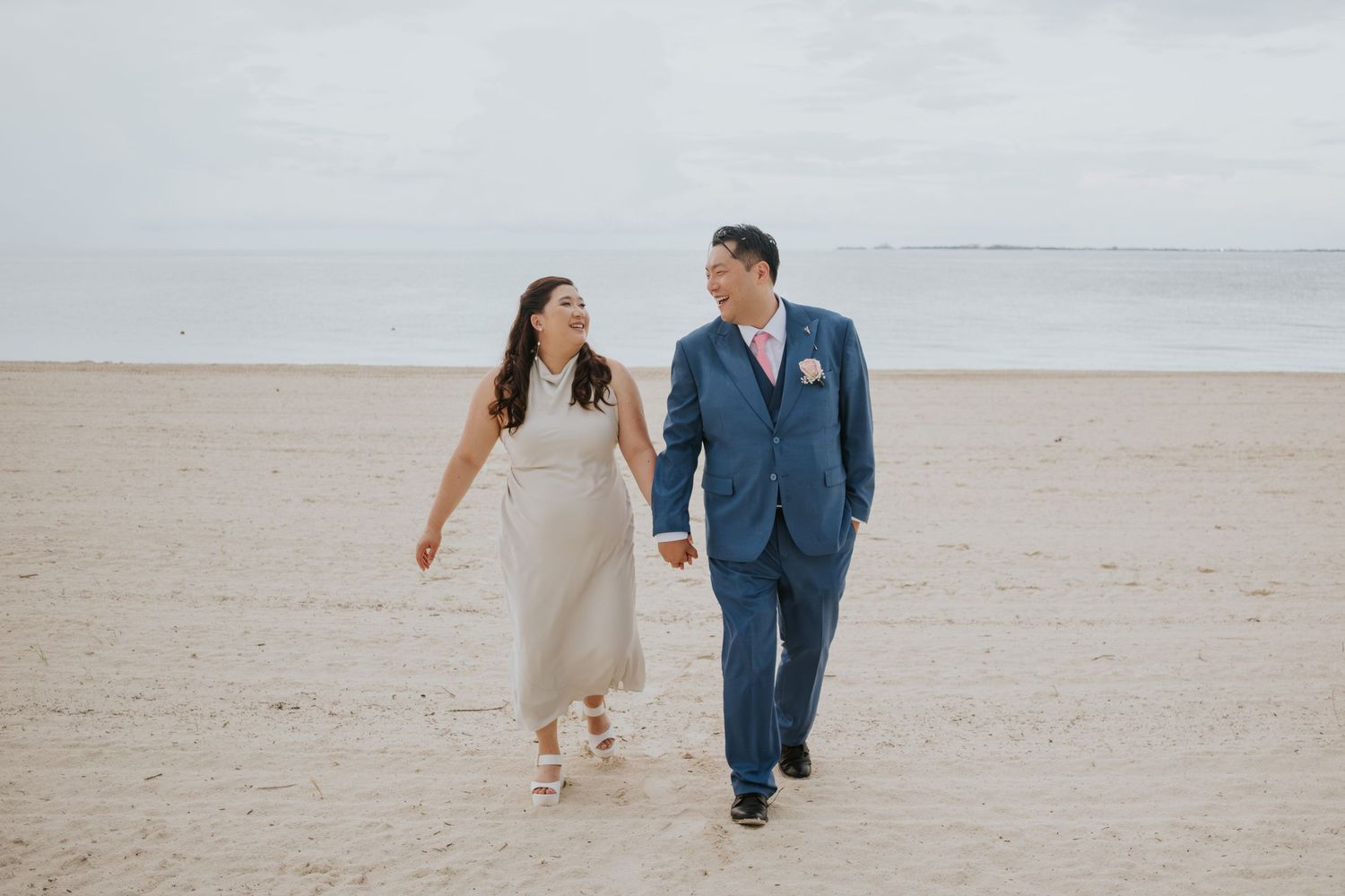 Newlywed couple walking hand in hand on a sandy beach, bride in white dress, groom in blue suit.