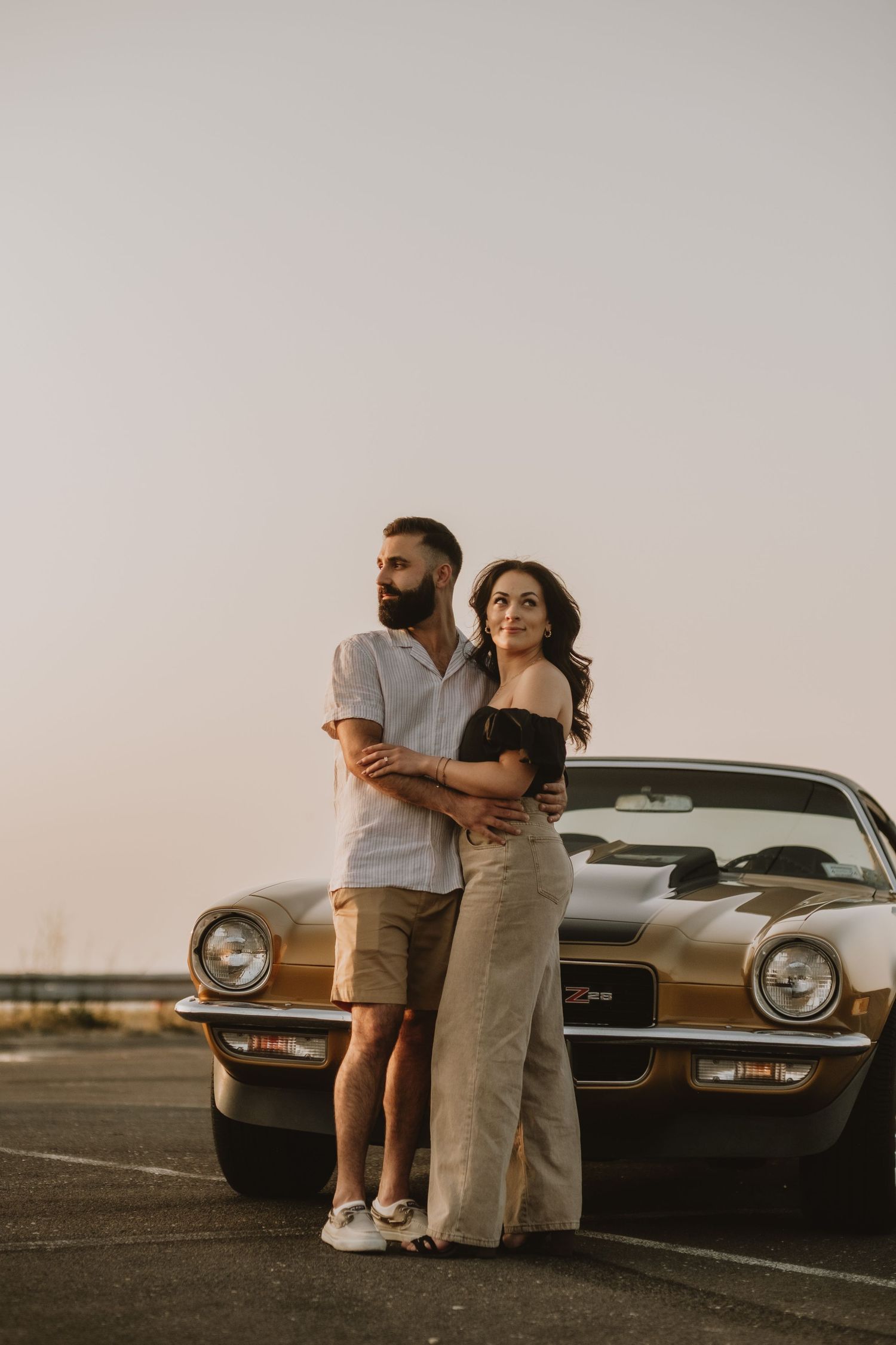 A couple poses together against a vintage Mustang car during golden hour in a parking lot.