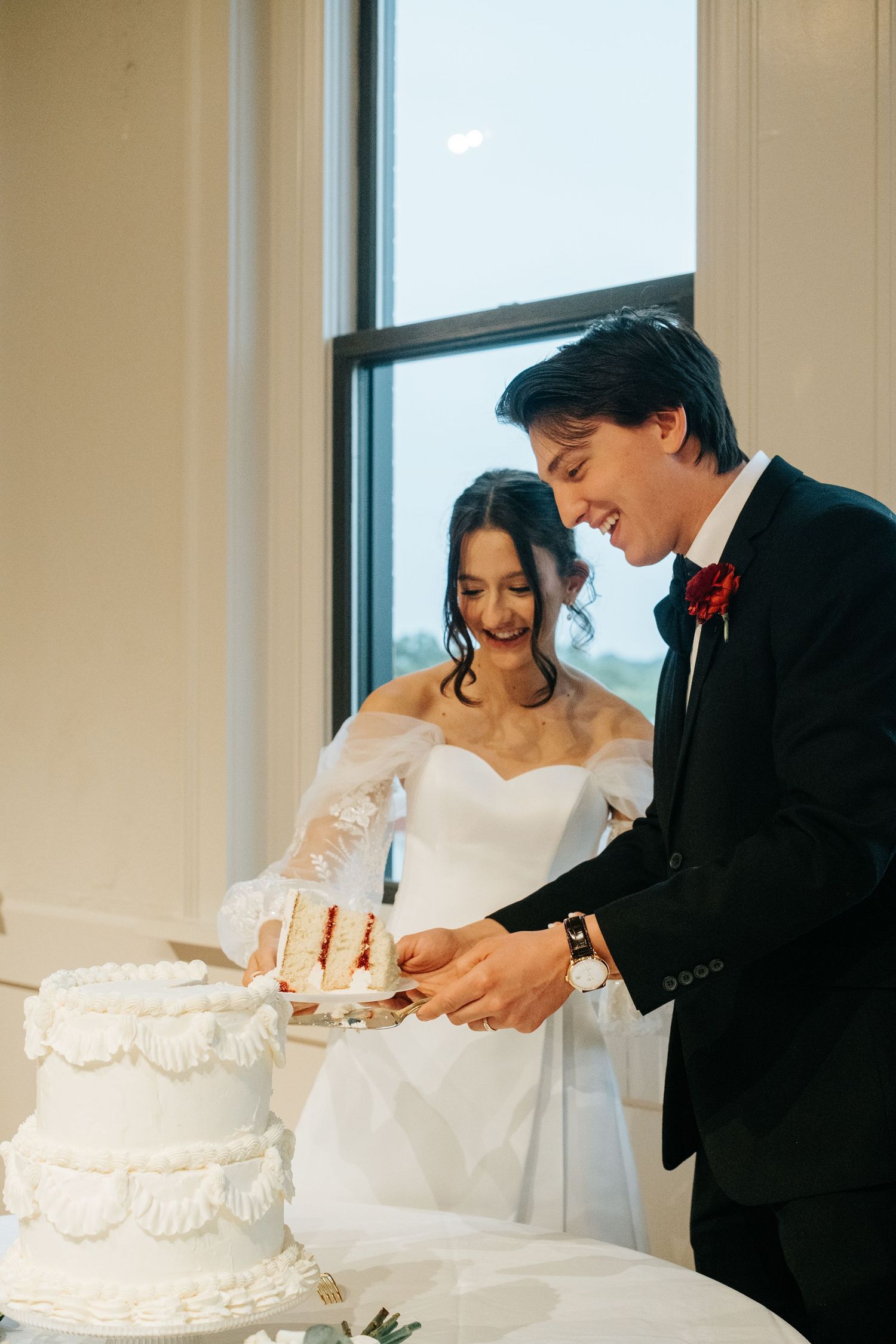 A couple cutting their elegant white tiered wedding cake together by a window.