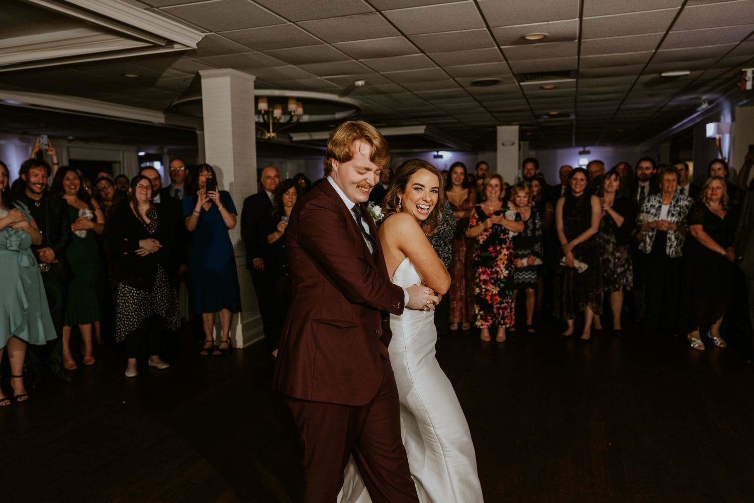 Bride and groom share first dance at wedding reception with guests clapping in background.