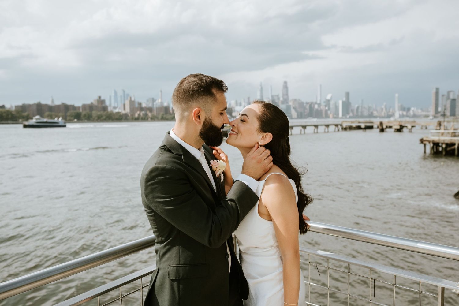 Bride and groom share a romantic moment by the waterfront with a city skyline backdrop on their wedding day.