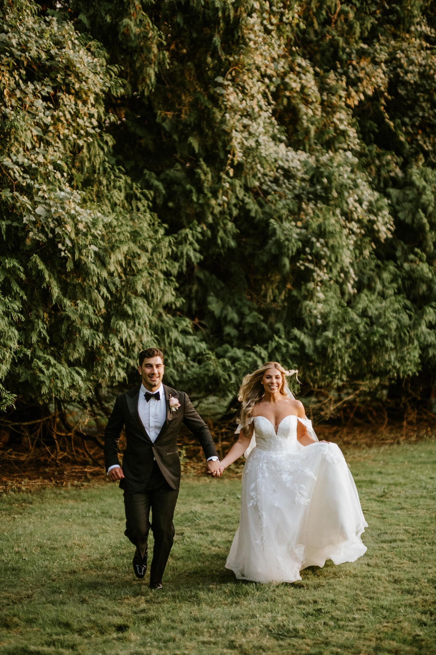 Joyful bride and groom running hand in hand across green lawn with lush trees in background on wedding day.