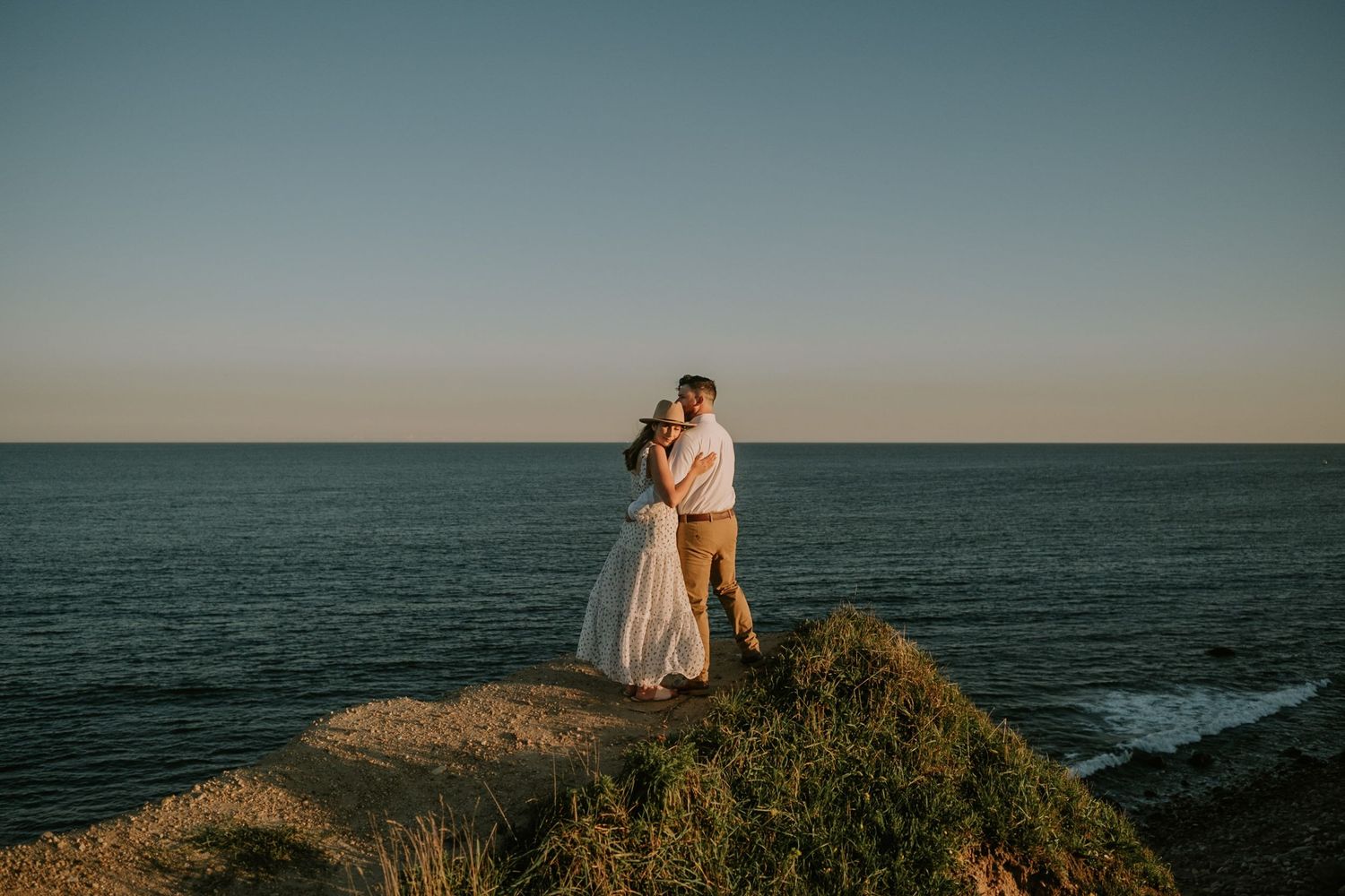 A couple embraces on a scenic coastal cliff overlooking the ocean at sunset.