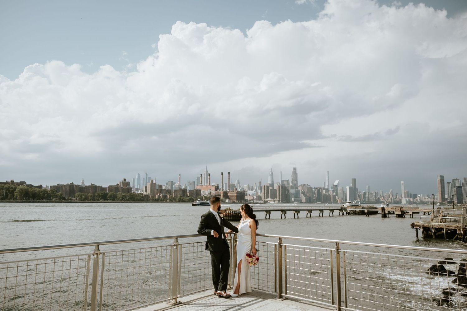 Couple exchanging vows on a waterfront pier with NYC skyline backdrop under dramatic cloudy skies.