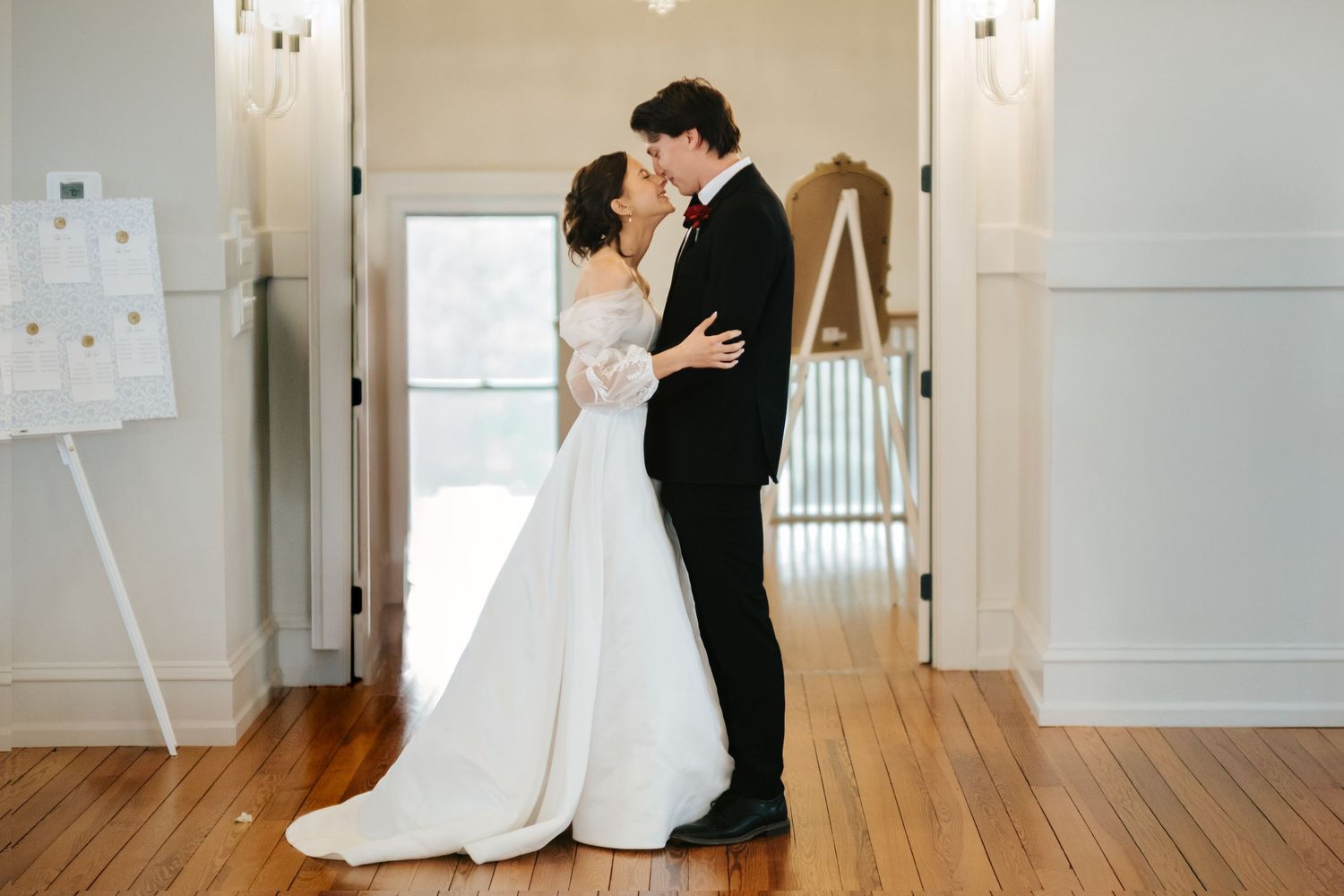 A romantic couple in formal wedding attire shares an intimate moment in an elegant white room with hardwood floors.