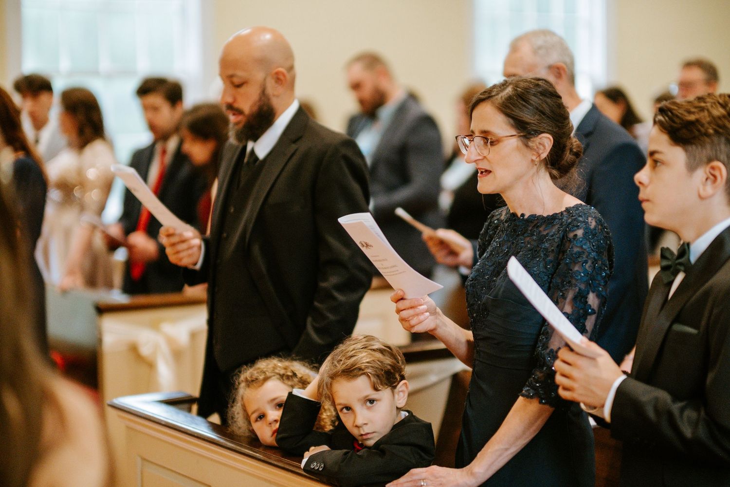 Wedding guests in formal attire reading from programs during a church ceremony service.