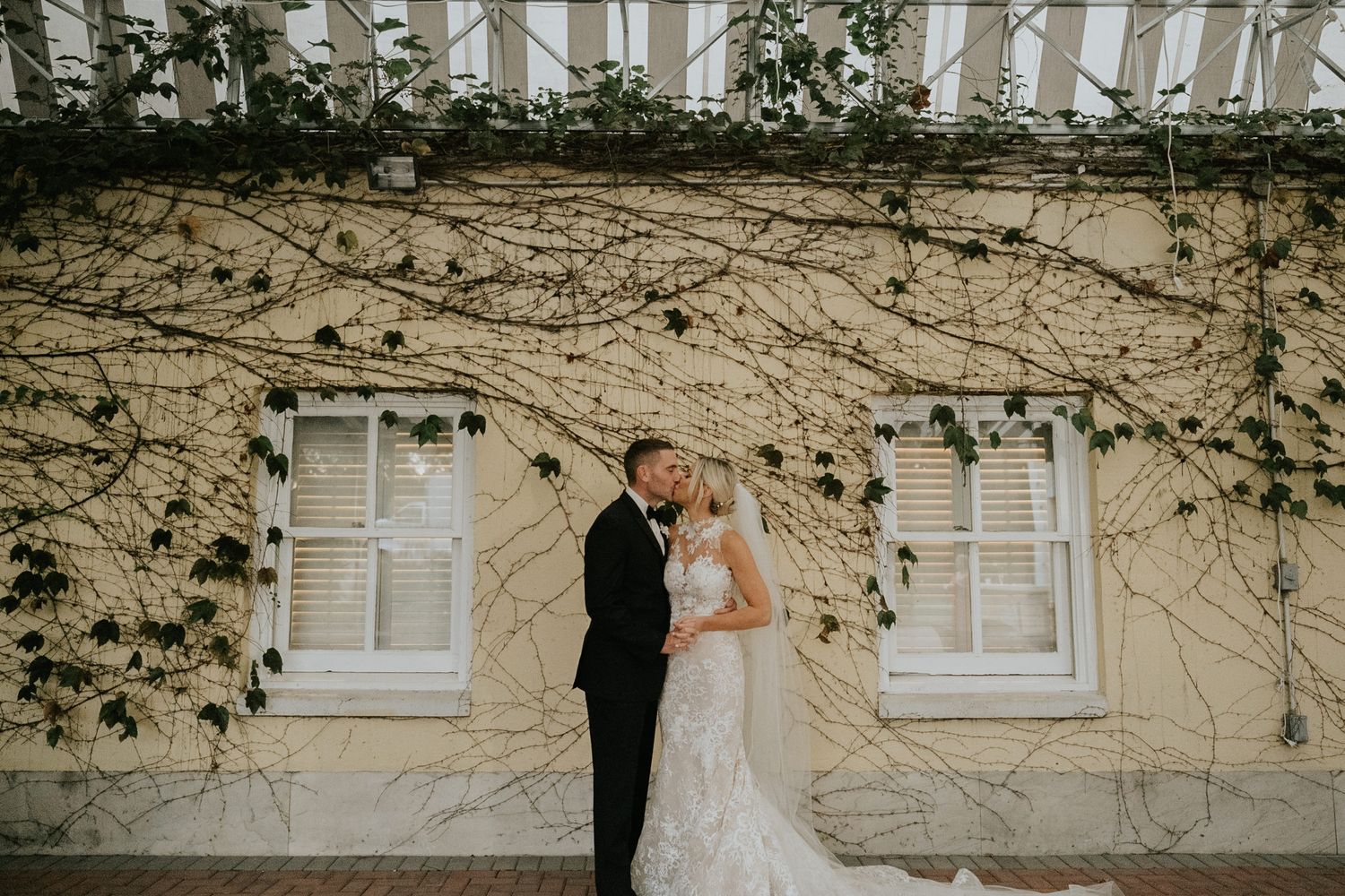 Bride and groom share a romantic kiss in front of a yellow vine-covered building with white windows.