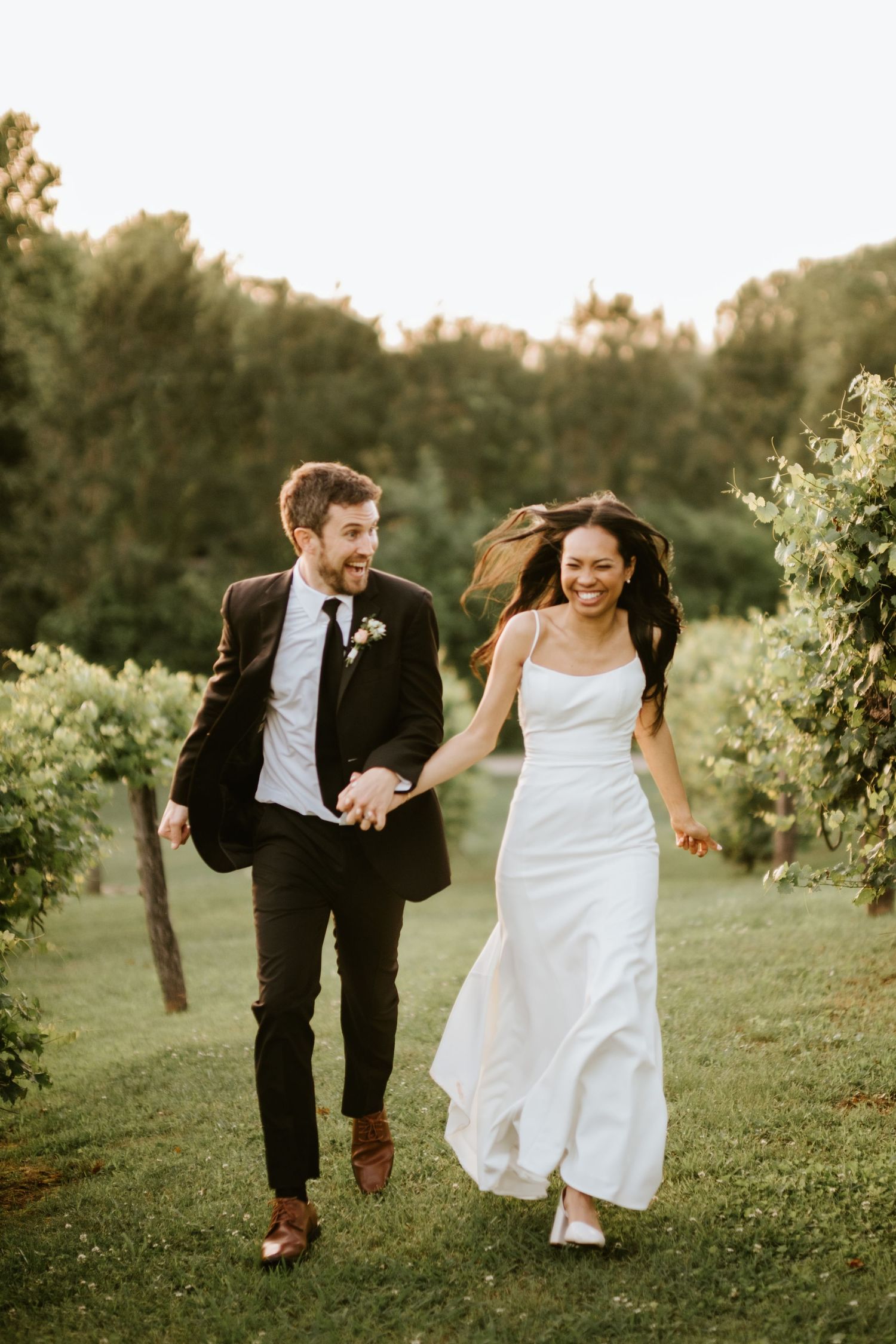 A couple in wedding attire runs playfully through a sunlit field at golden hour.