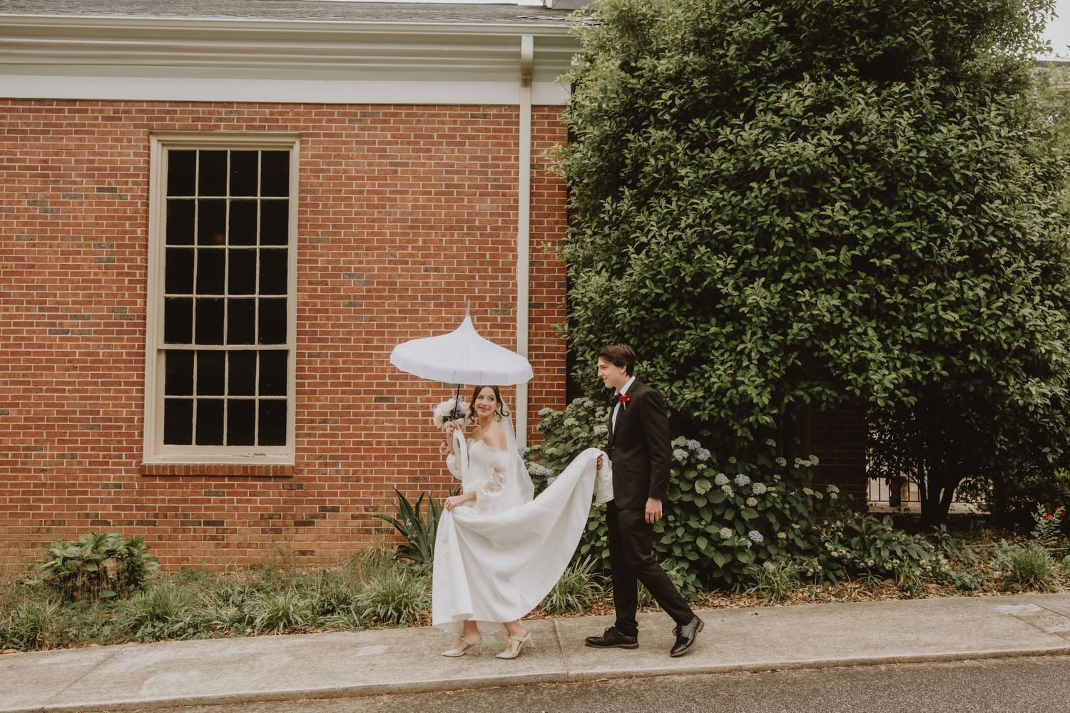 A romantic wedding couple walks along a brick building exterior with white columns and windows.