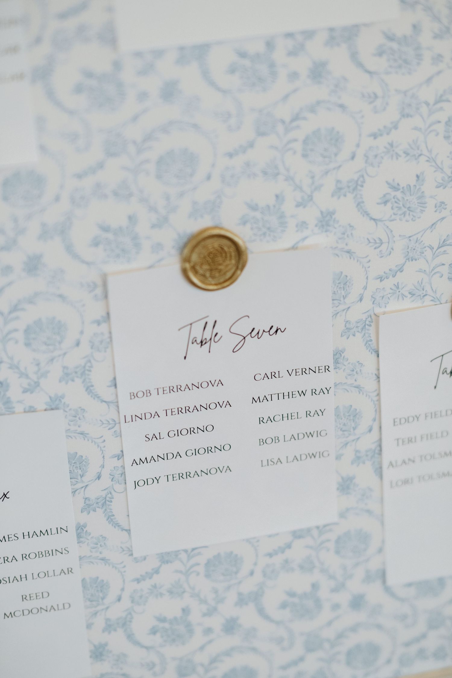 Wedding table seating chart with gold wax seal displayed on light blue floral patterned background.