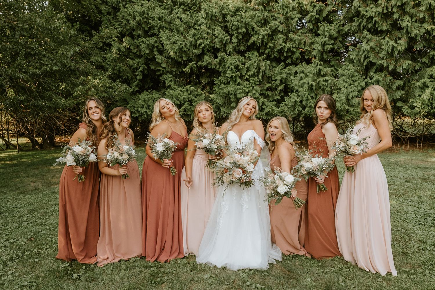 Bride in white gown poses with seven bridesmaids in rust and blush dresses holding bouquets outdoors.