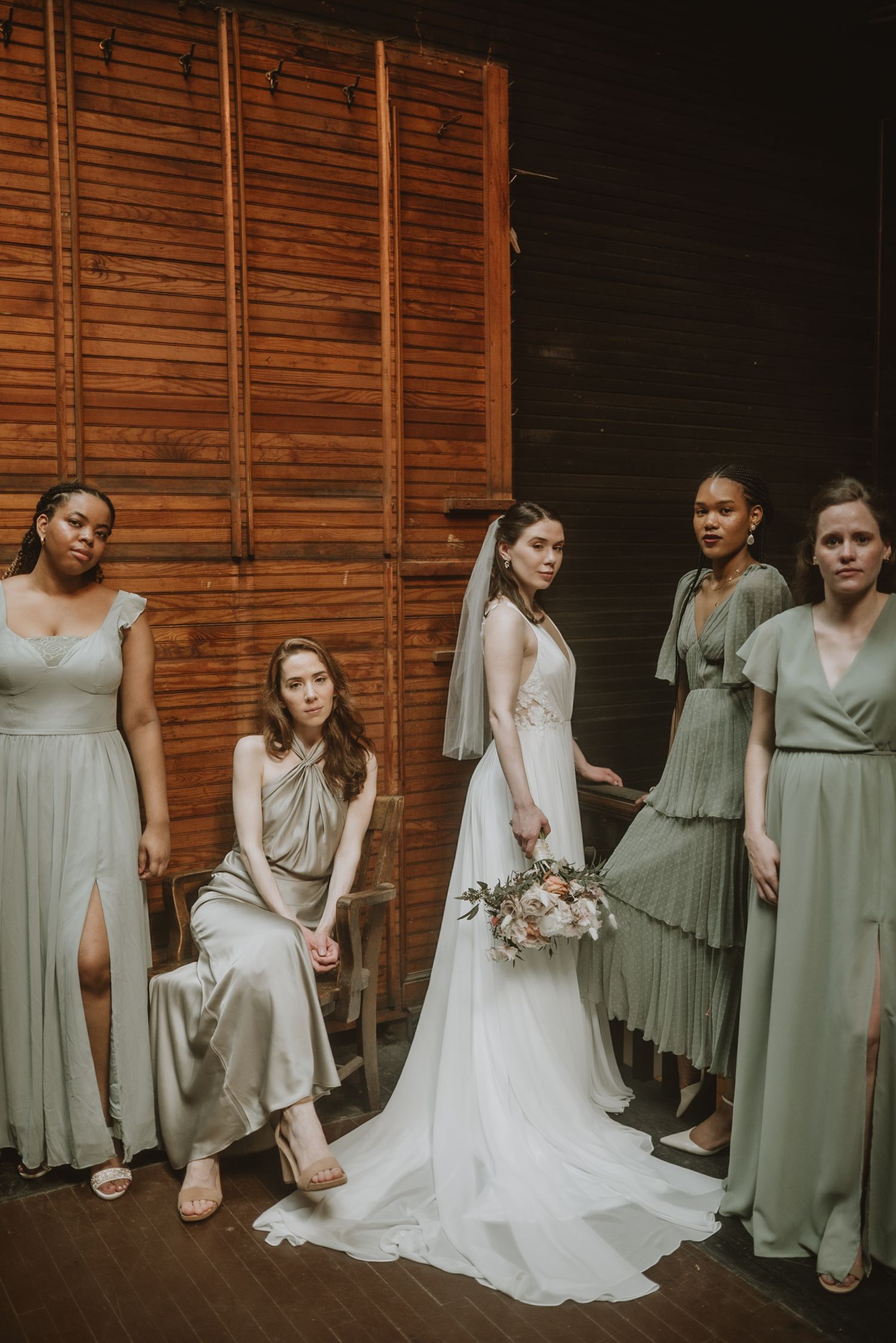 Bride in white gown with bouquet poses with four bridesmaids in sage green dresses inside a rustic barn venue.