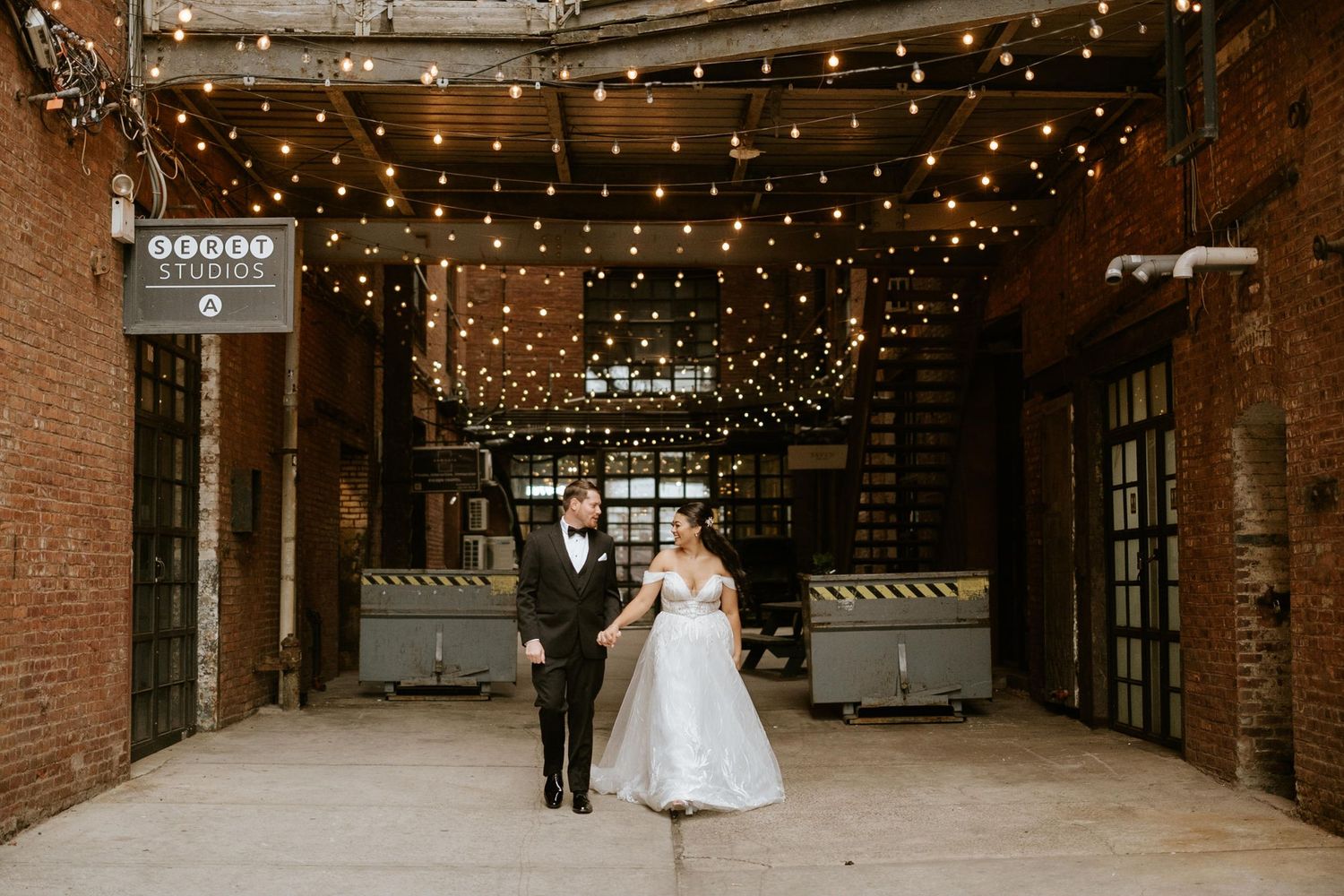 Bride and groom hold hands under string lights in a rustic brick venue alley during their wedding celebration.