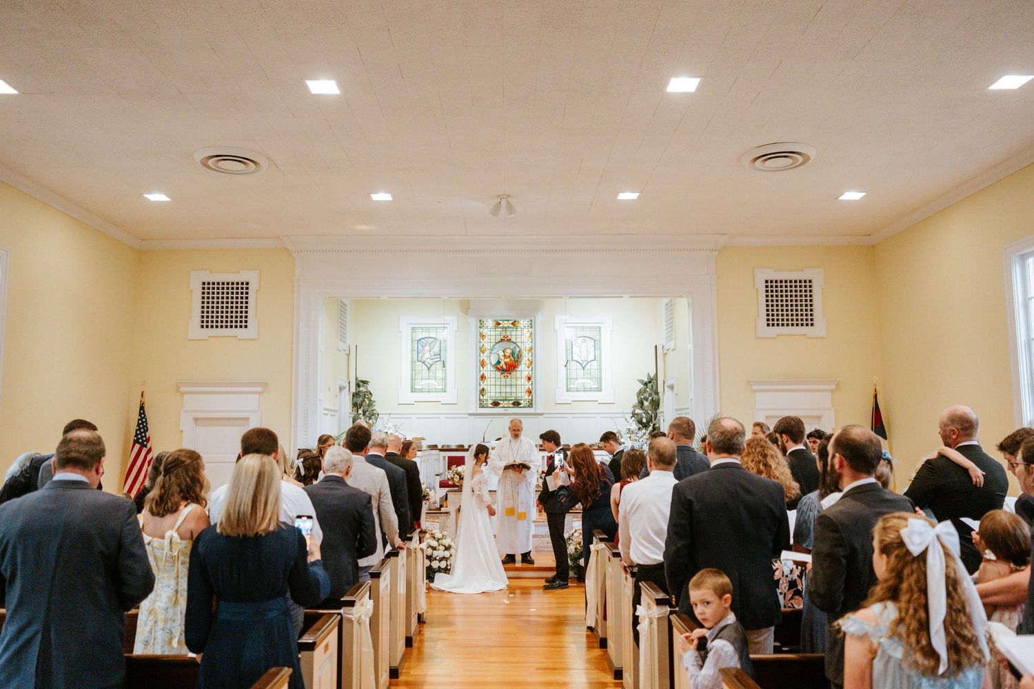 A wedding ceremony taking place in a bright chapel with wooden floors and white walls as guests observe from the pews.