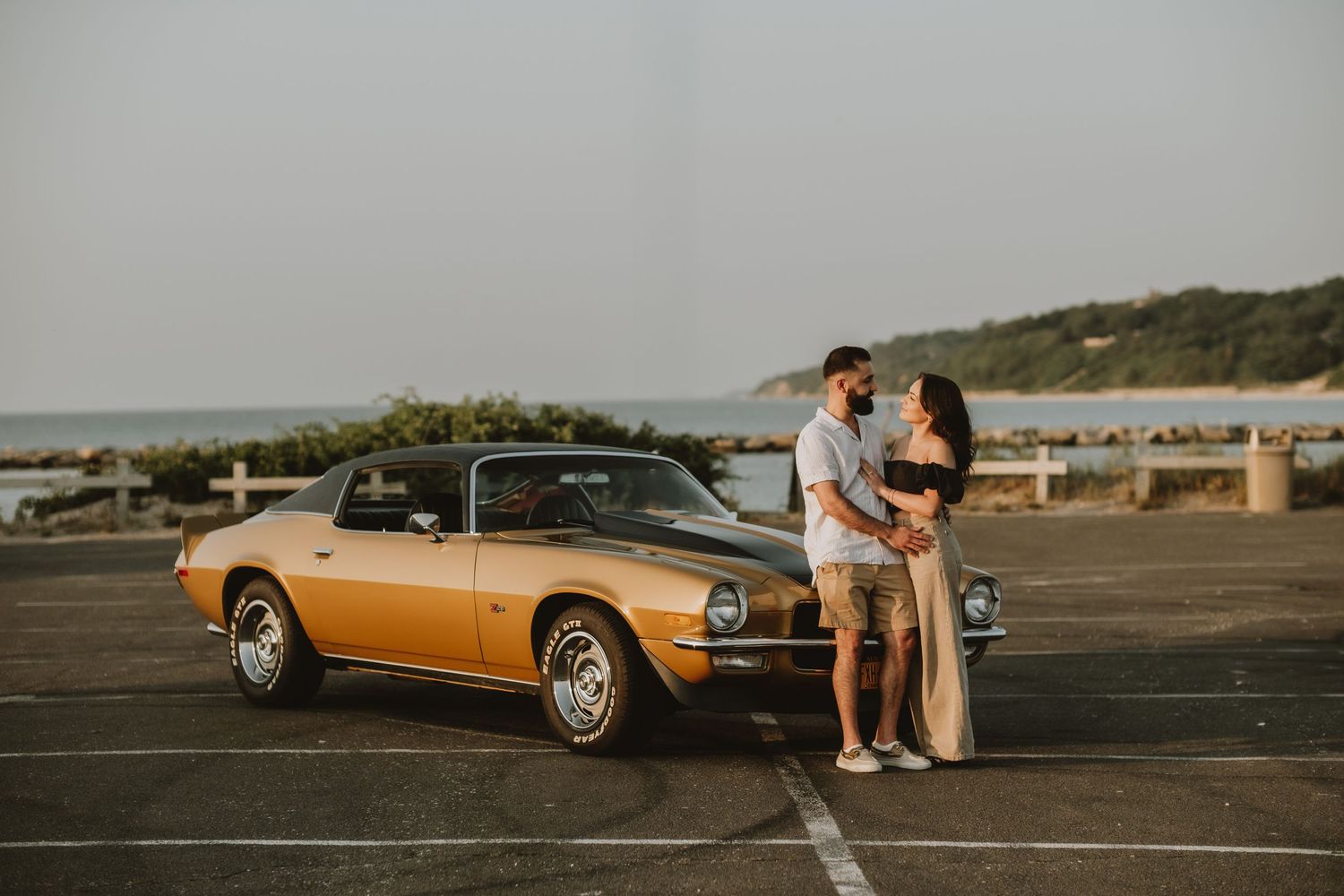 A classic gold convertible car parked at sunset on a coastal road as a couple sits on the hood watching the view.