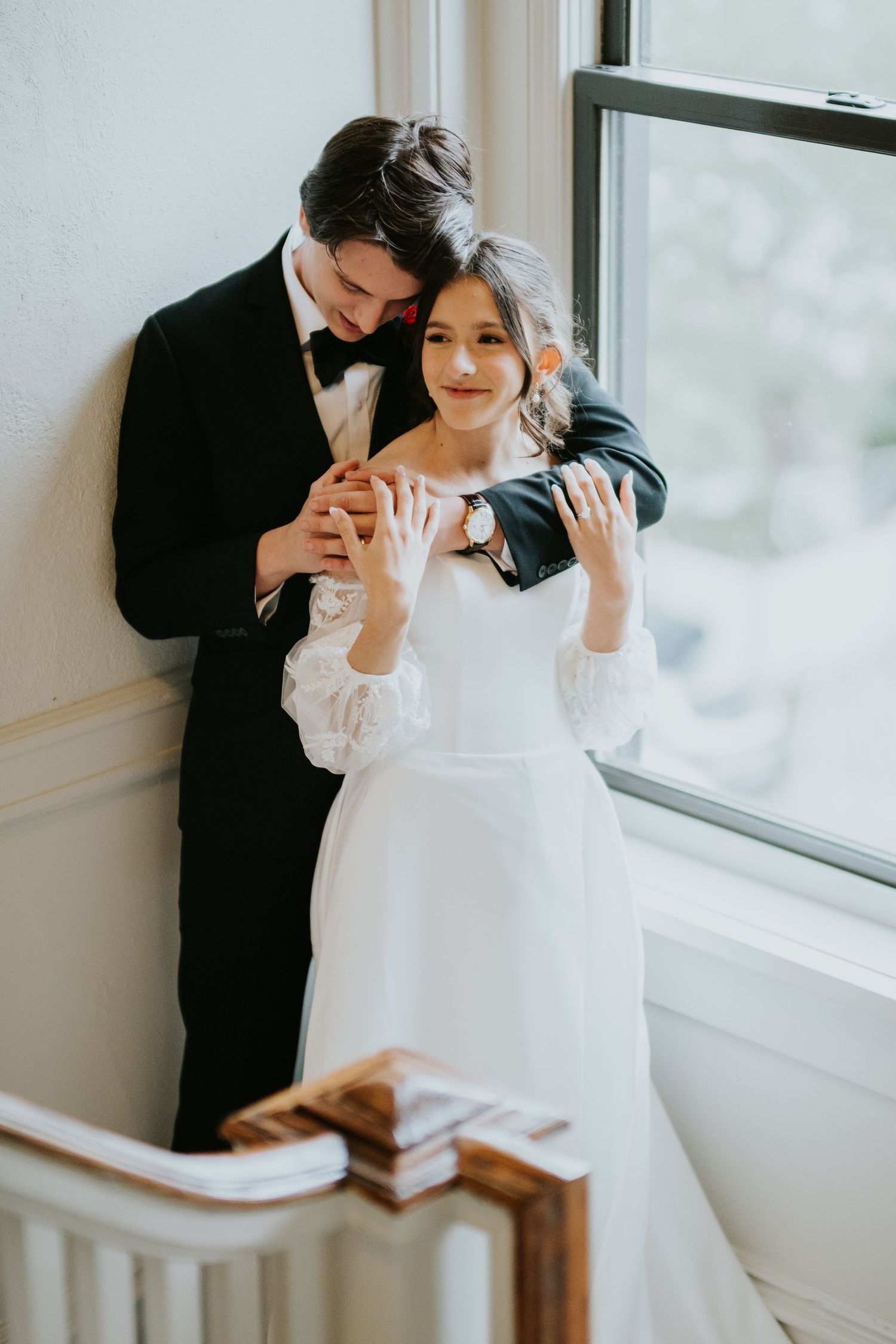 A couple shares a joyful embrace near a window, with the person in white wearing long sleeves.