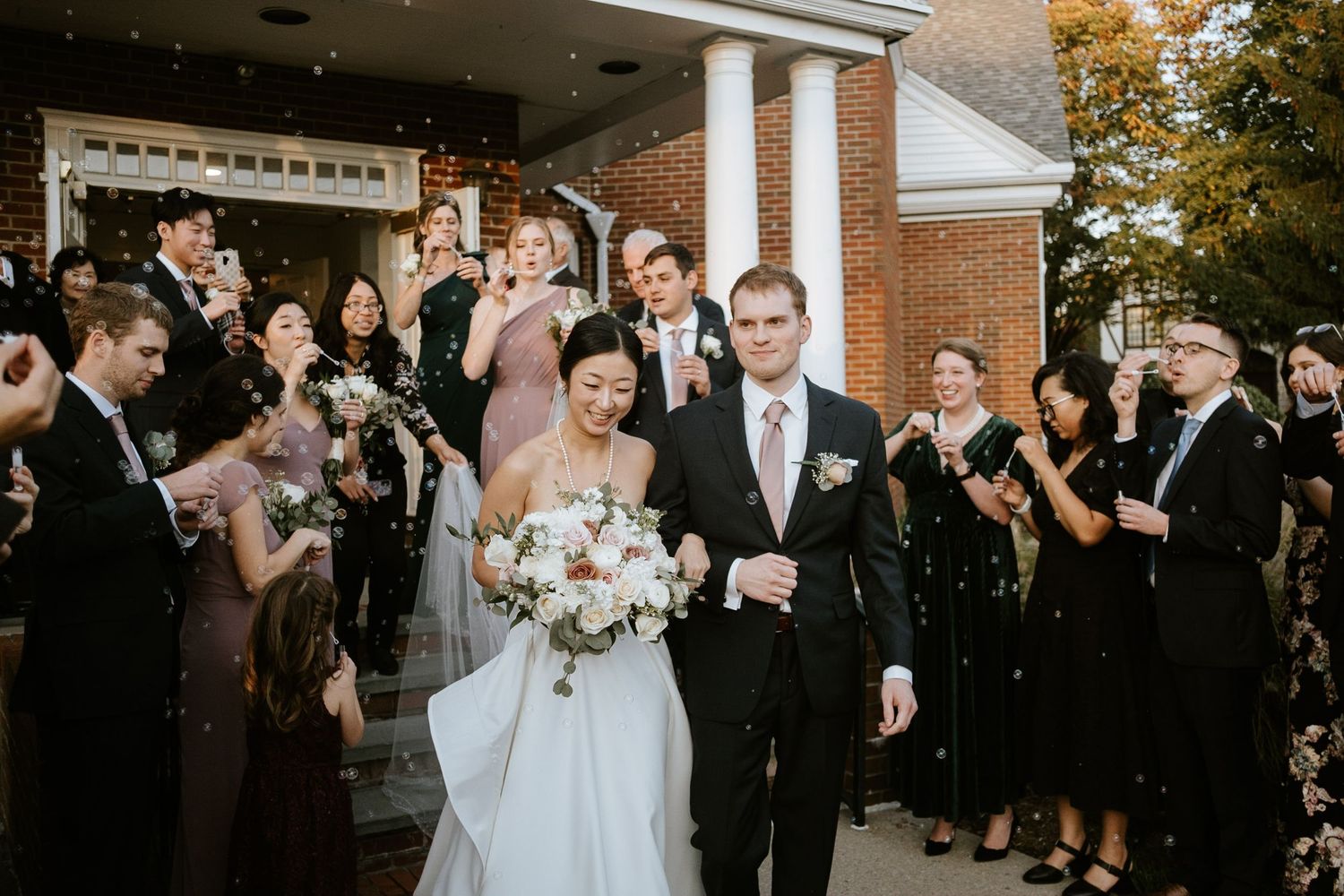 Bride and groom exit wedding venue as guests toss confetti, bride holding white floral bouquet.