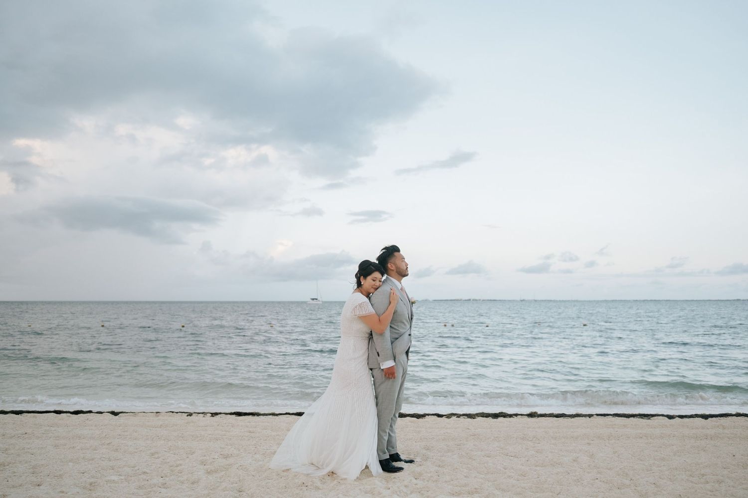 Bride and groom embrace on a sandy beach at dusk with calm ocean waves and cloudy sky in the background.