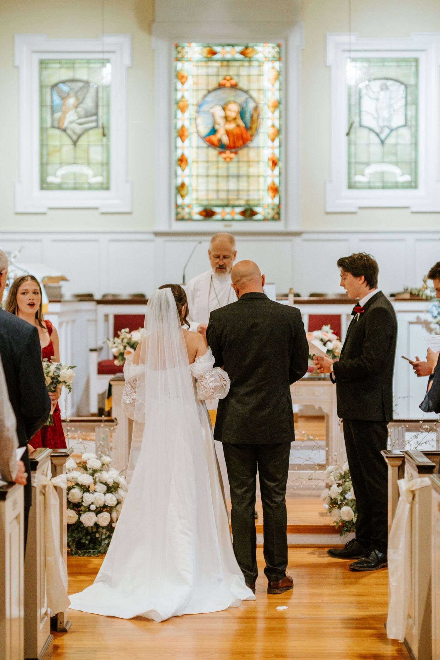A wedding ceremony takes place in a beautiful church with stained glass windows and floral decorations along the aisle.