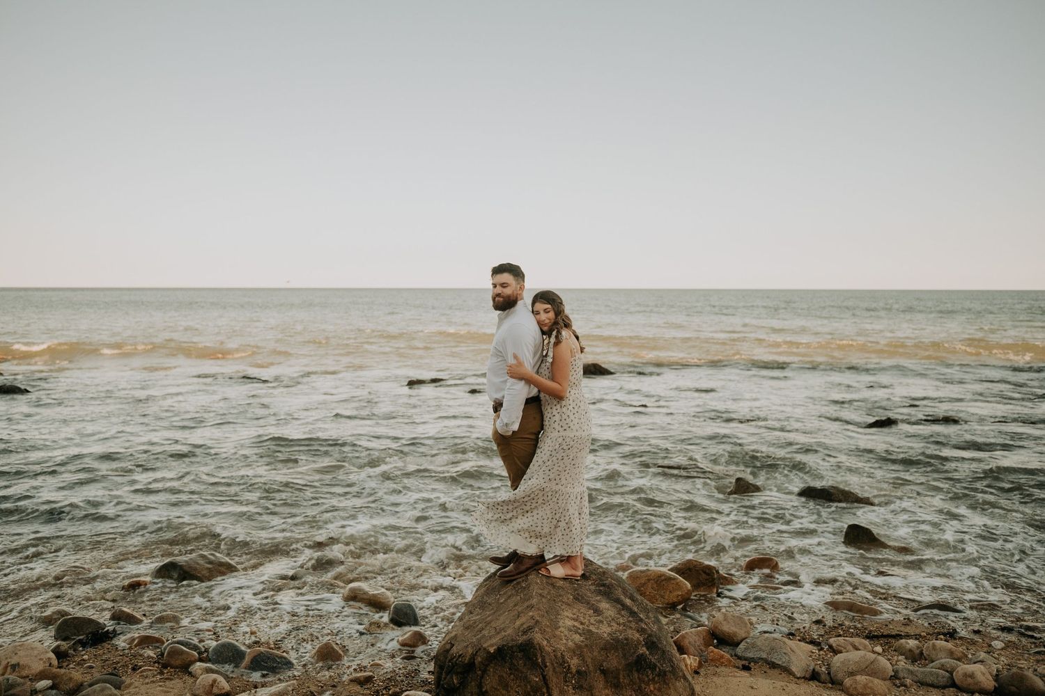 Couple embracing on rocky ocean shore at sunset, woman in lace dress and man in white shirt, waves behind them.