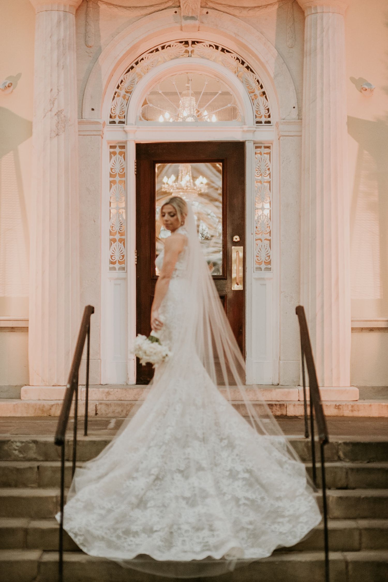 Bride in lace mermaid gown with cathedral veil stands on steps of elegant white columned venue holding white bouquet.